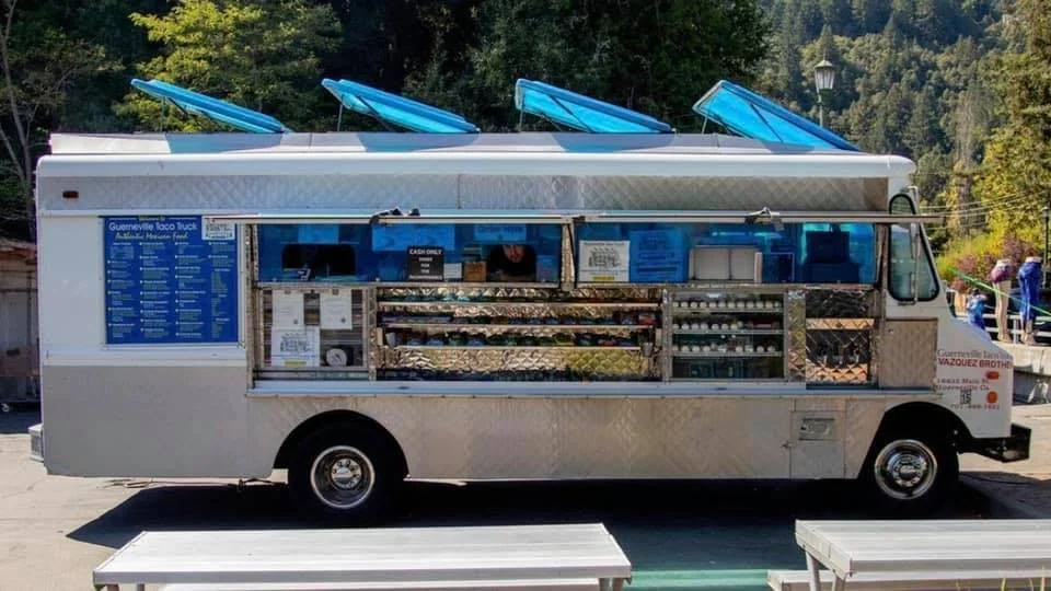A mobile food truck with blue umbrellas on the roof, serving Mexican food, parked outdoors with a backdrop of trees and mountains.