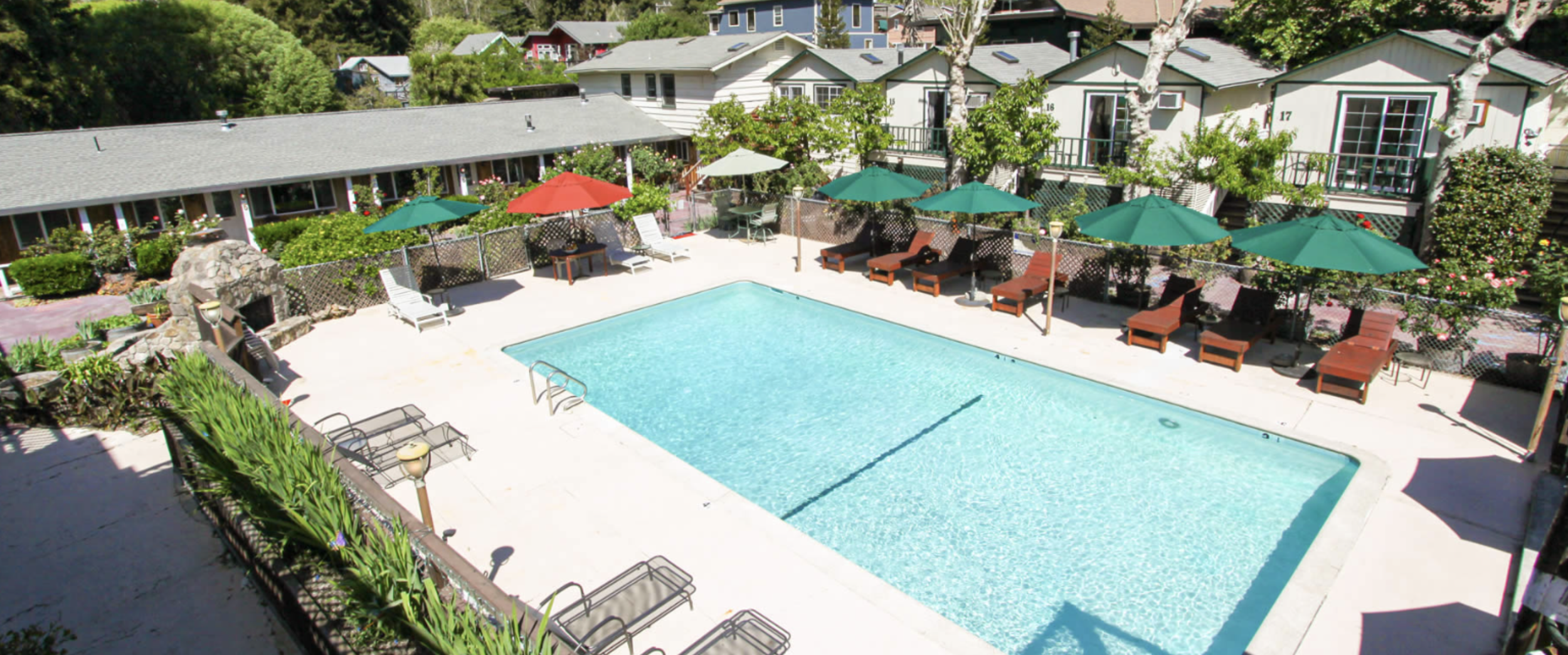 A swimming pool area with lounge chairs and colorful umbrellas, surrounded by greenery and residential buildings.