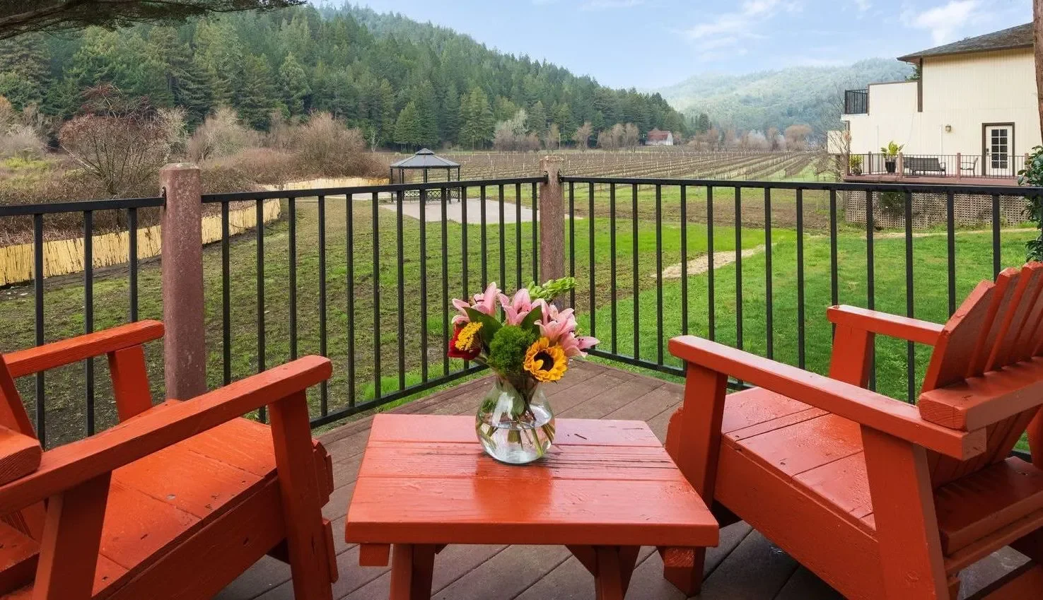 A backyard patio with two orange wooden chairs and a small matching table with a flower vase, overlooking a green grassy yard, a black metal fence, trees, hills, and a gazebo in the distance.
