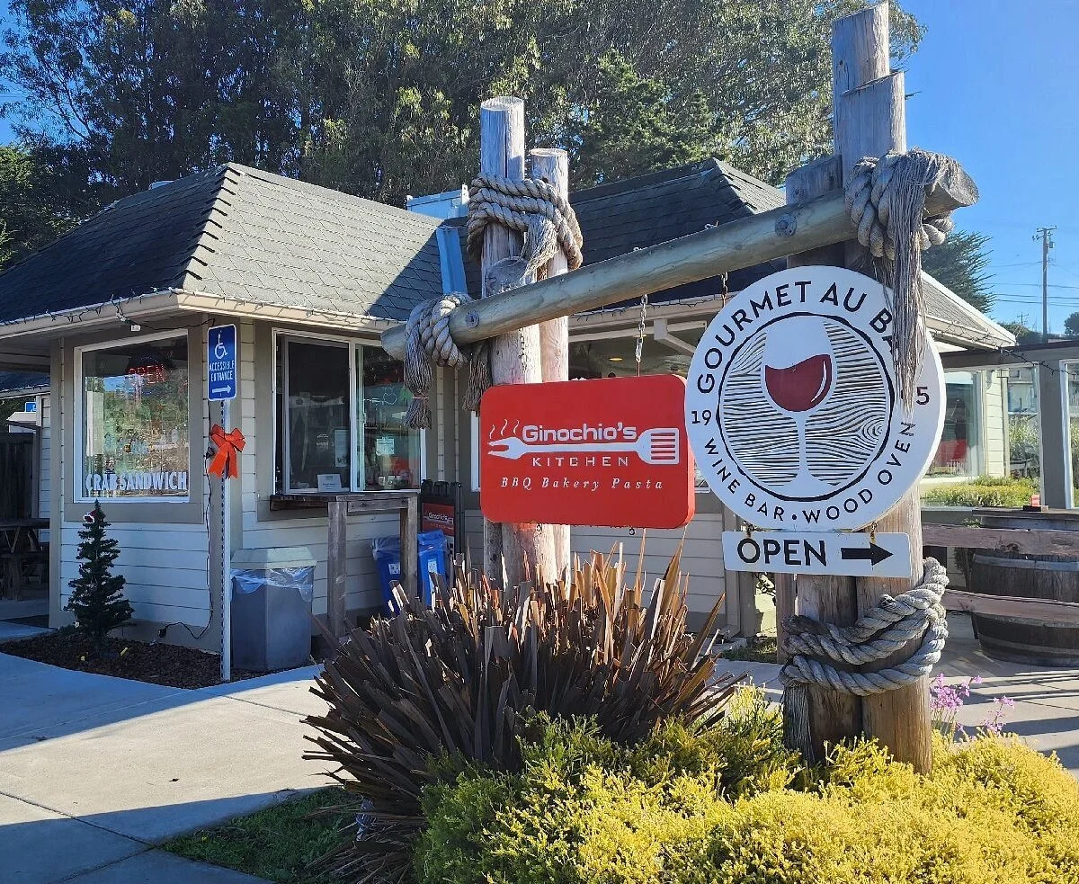 Signboard outside Ginochio's Kitchen, a restaurant offering BBQ, bakery, and pasta, with a wine bar and wood oven, located in a small building with a sidewalk and garden plants in front.