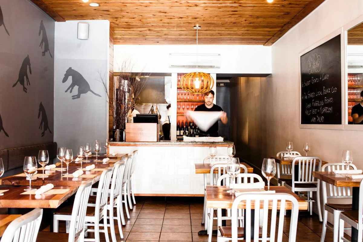 Restaurant dining area with wooden tables, white chairs, wine glasses set on tables, and a man preparing food behind the counter.