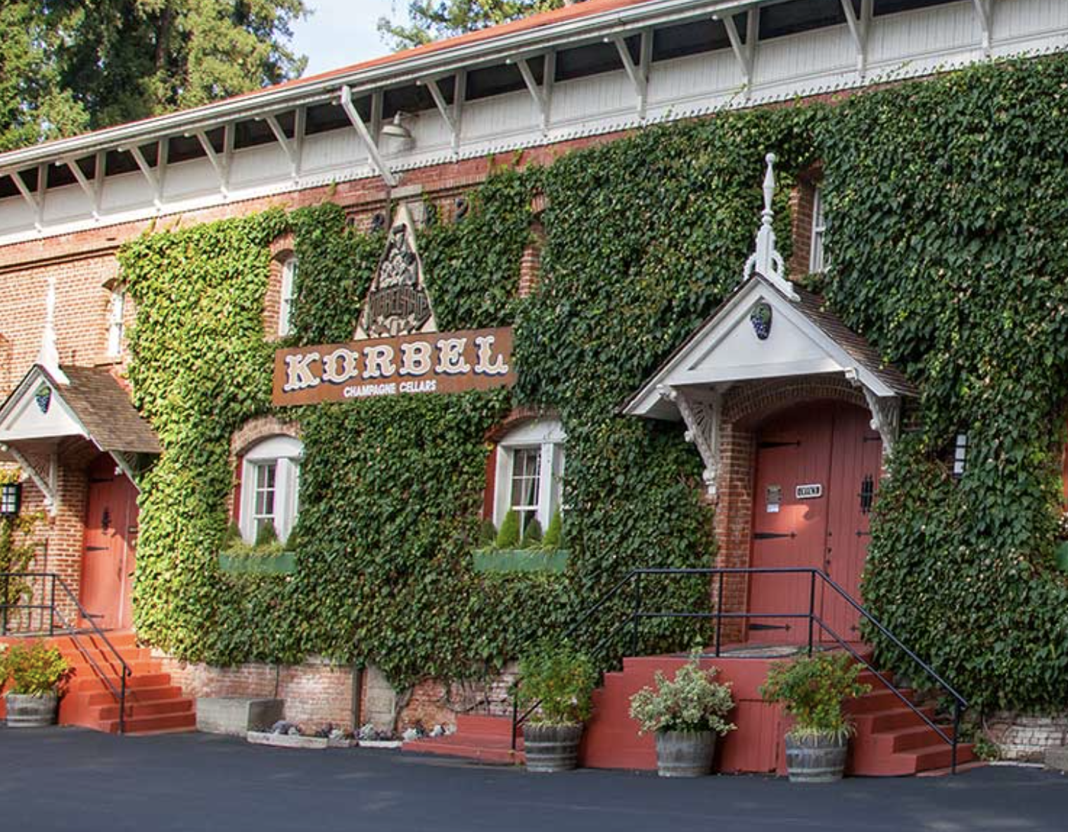 Building covered with green ivy, with a sign that says 'Korbel Champagne Cellars,' featuring a red door and steps, and an ornate white porch roof.