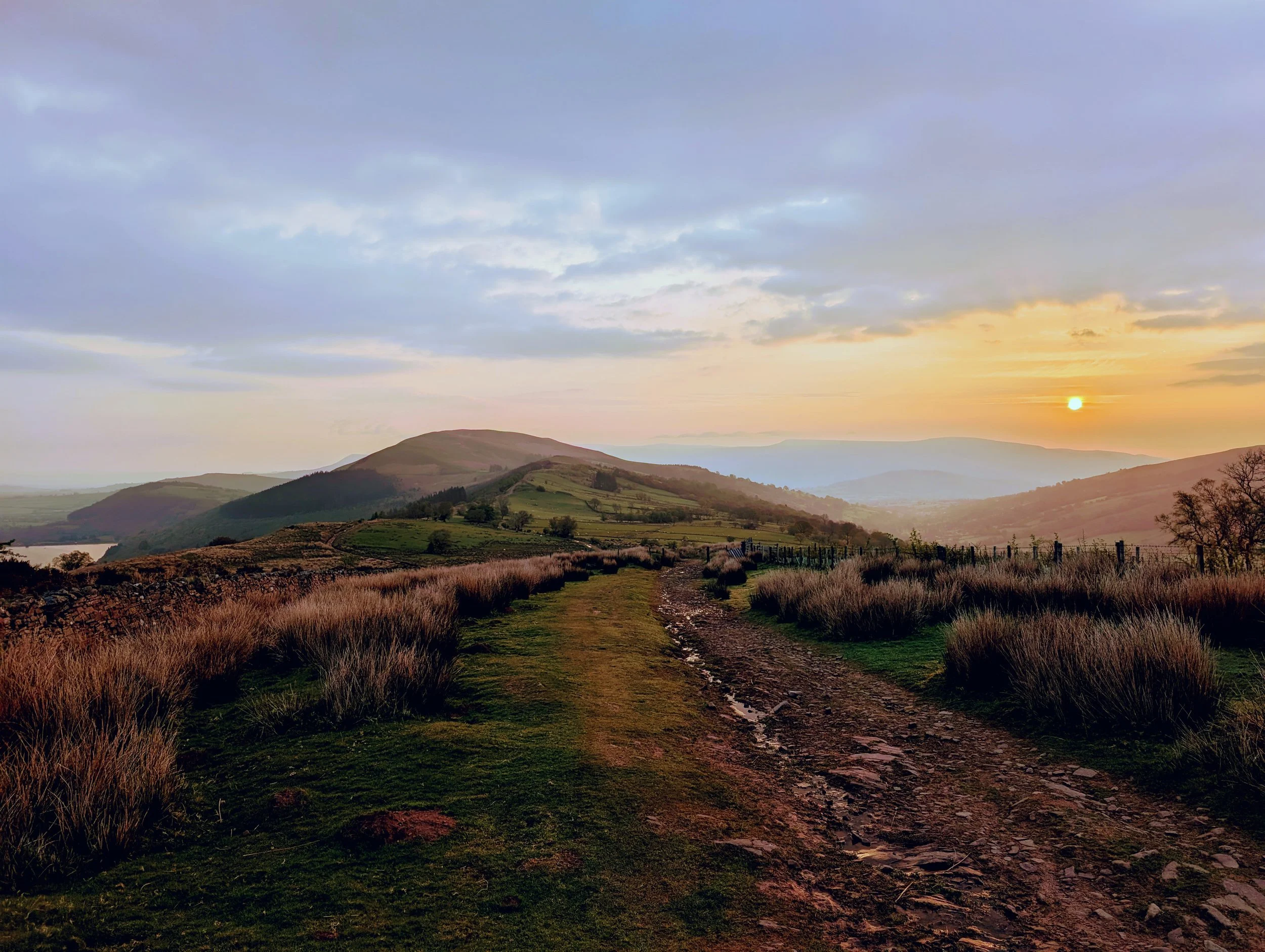 A landscape scene at sunset showing rolling hills, a dirt path, and a partly cloudy sky with the sun near the horizon.