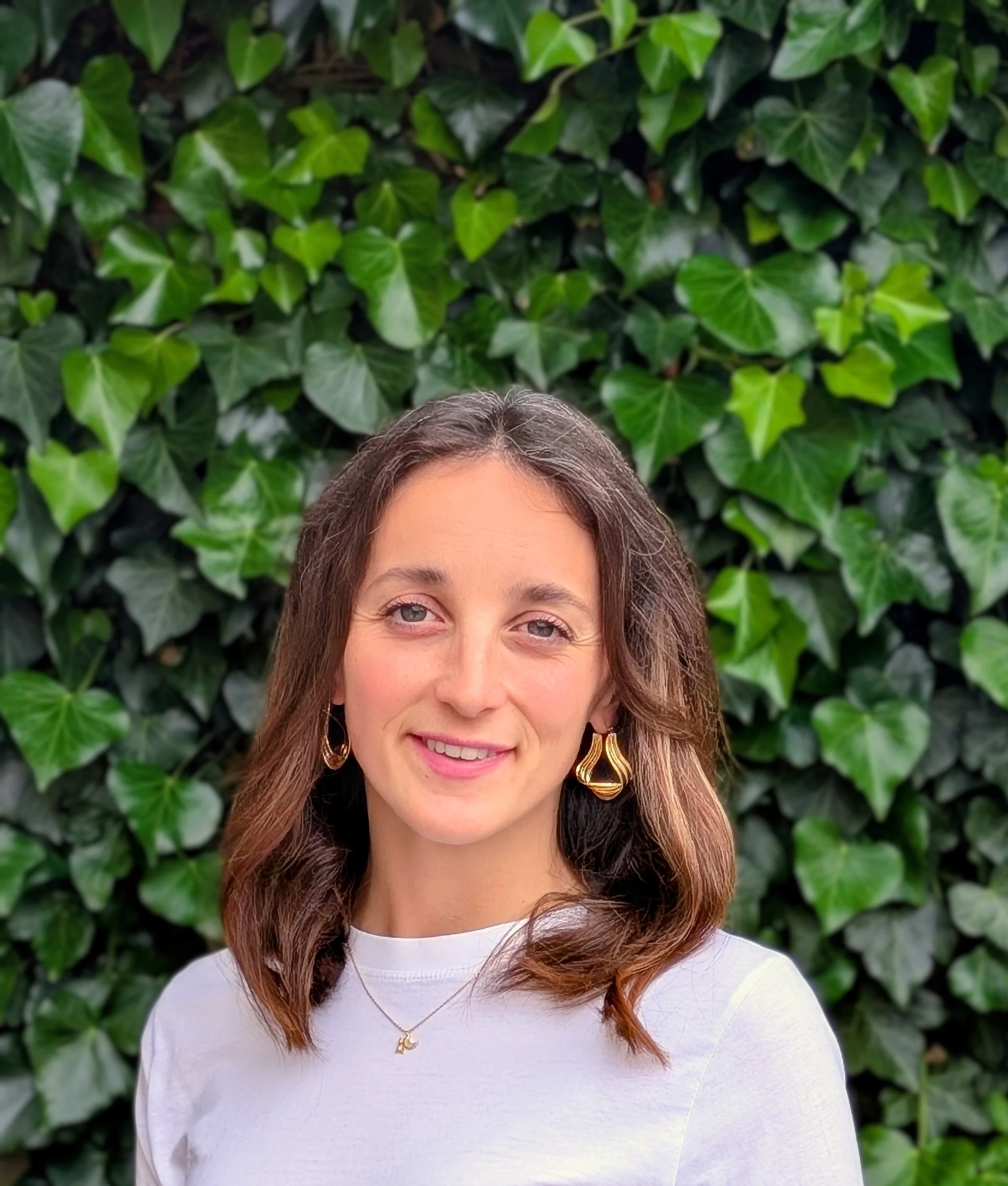 A female counsellor with brown hair wearing a white top, gold earrings, and a gold necklace with a moon pendant, standing in front of a green leafy background.