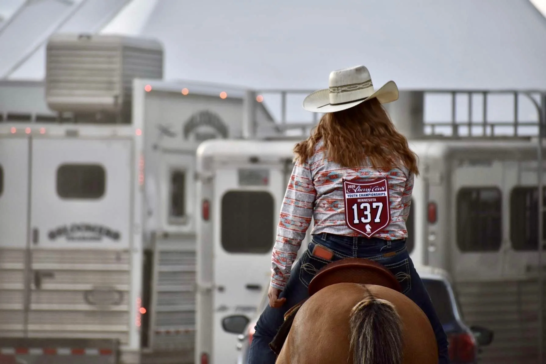 A person wearing a cowboy hat and a rodeo competition shirt riding a horse in front of horse trailers at a rodeo event.