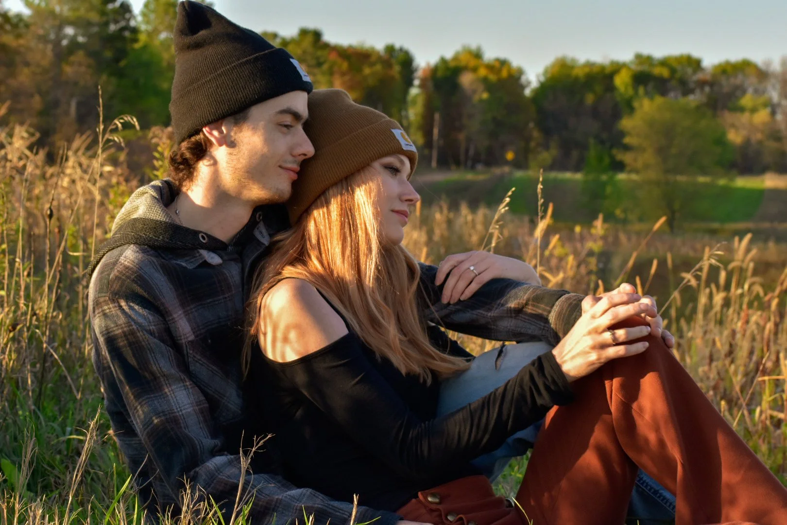 A young couple sitting together in a grassy field during sunset, embracing each other and enjoying a peaceful outdoor moment.