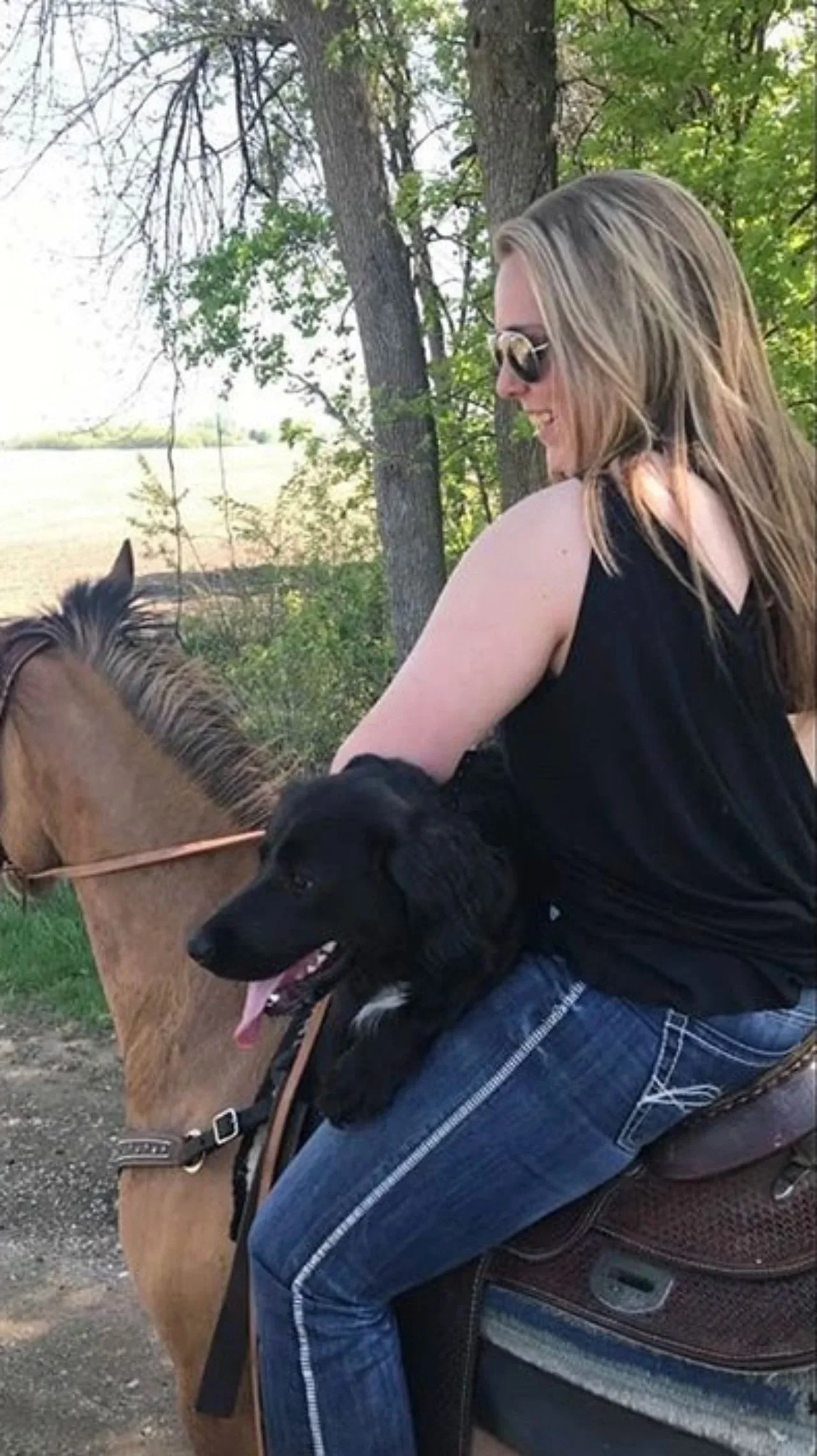 A woman with long blonde hair, wearing sunglasses, a black sleeveless top, and blue jeans, is sitting on a horse outdoors. She is smiling and holding a black dog, which is sitting in her lap. The background features trees and a bright sky.