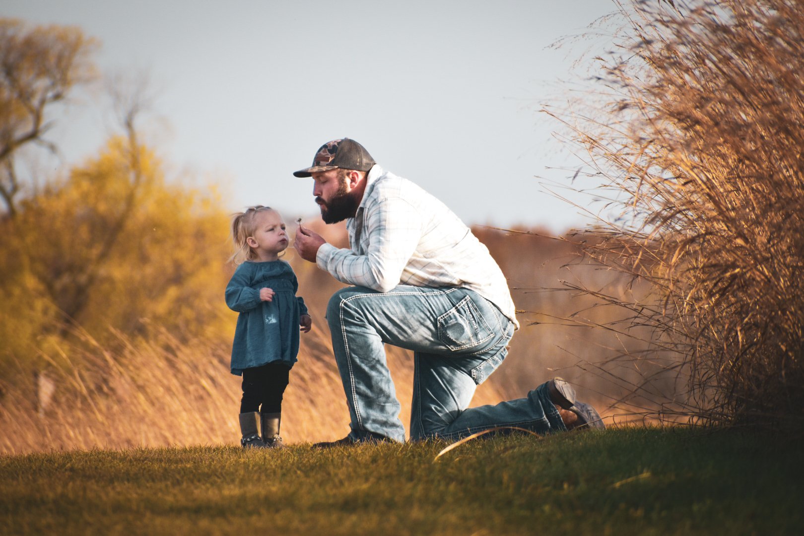 A man kneeling outdoors on grassy ground, blowing on a dandelion with a young girl standing facing him, with a background of tall, golden grass and trees with fall foliage.