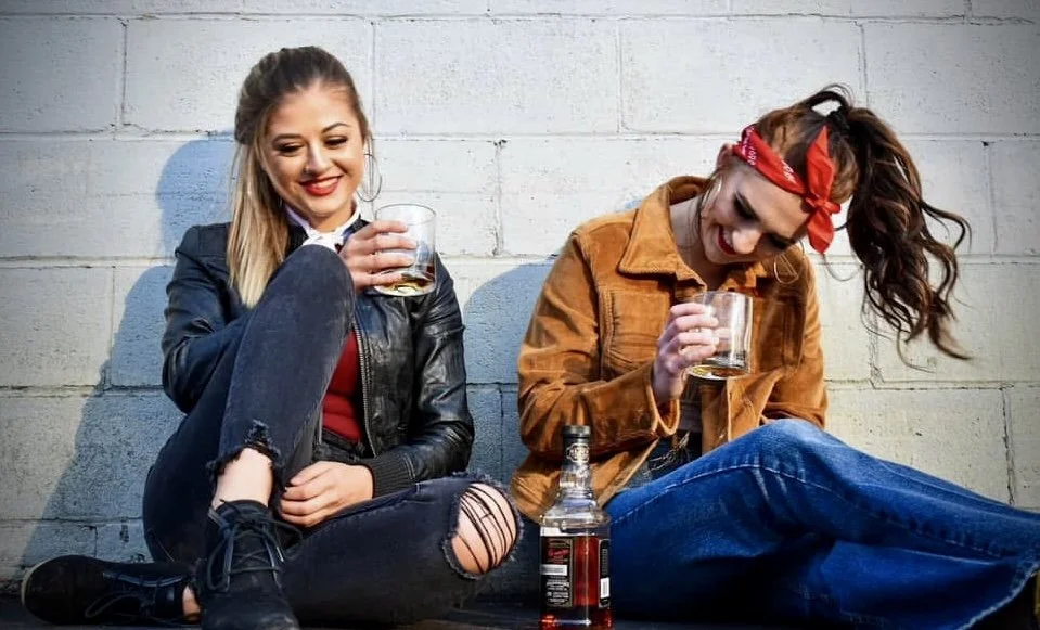 Two women sitting against a white brick wall, holding glasses of whiskey, with a whiskey bottle on the ground between them.