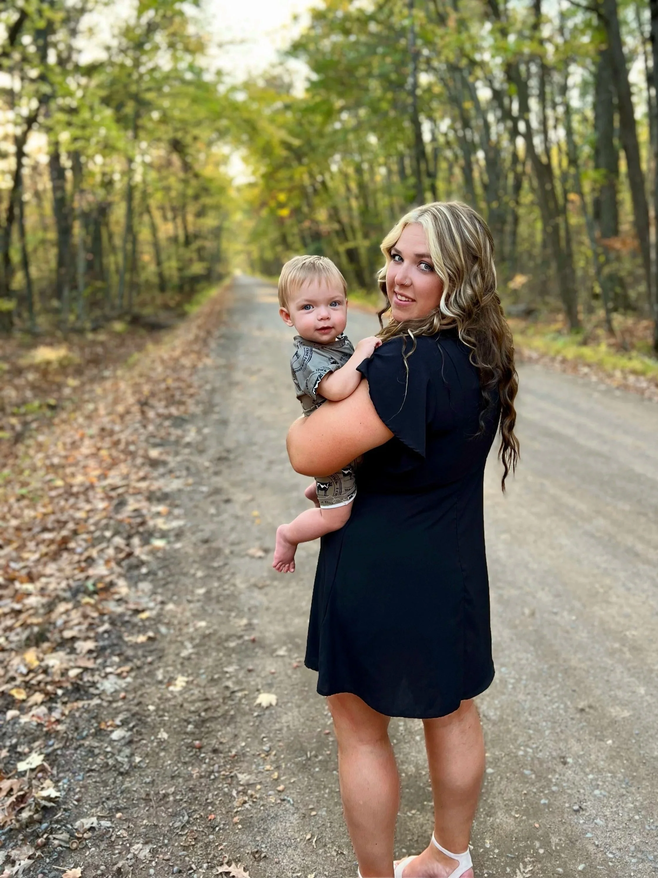 A woman with long, wavy blonde hair, wearing a knee-length black dress, holding a young boy with short blonde hair, wearing a grey patterned shirt and shorts, standing on a dirt path in a wooded area with green and yellow leaves.
