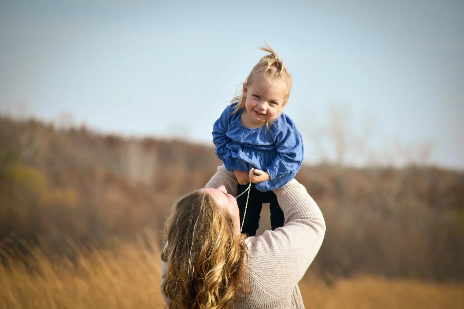 A woman lifting a young girl in an outdoor field during daytime, with both smiling and enjoying the moment.