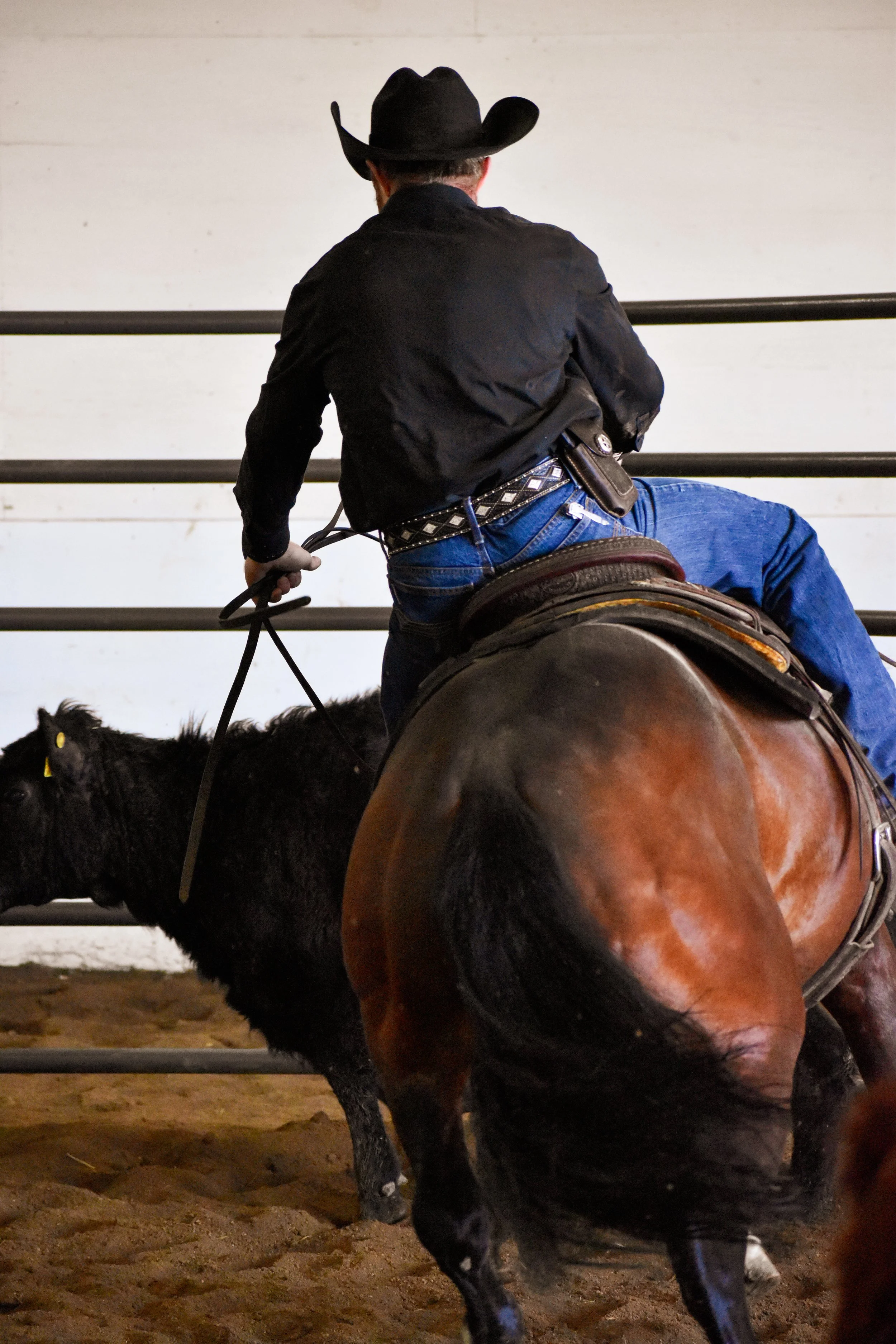 A man riding a horse inside an arena or rodeo, viewed from behind. The man is wearing a cowboy hat, a black shirt, jeans, and has a holstered gun on his belt. The horse has a brown saddle and is standing on dirt ground, with metal railings in the background.