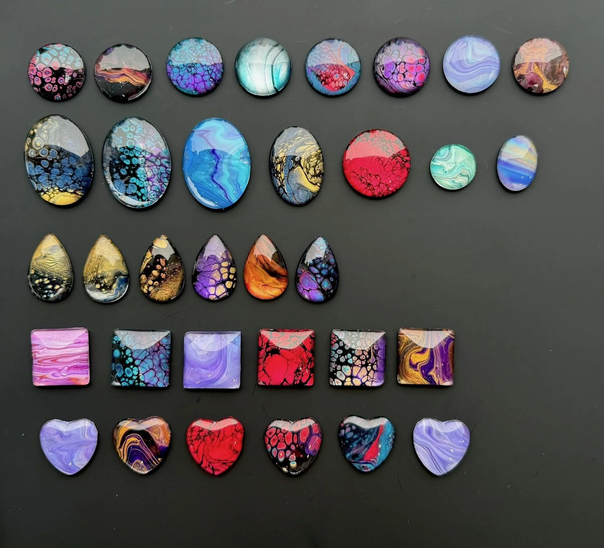 A collection of colorful, abstract glass or stone cabochons arranged on a dark gray background.