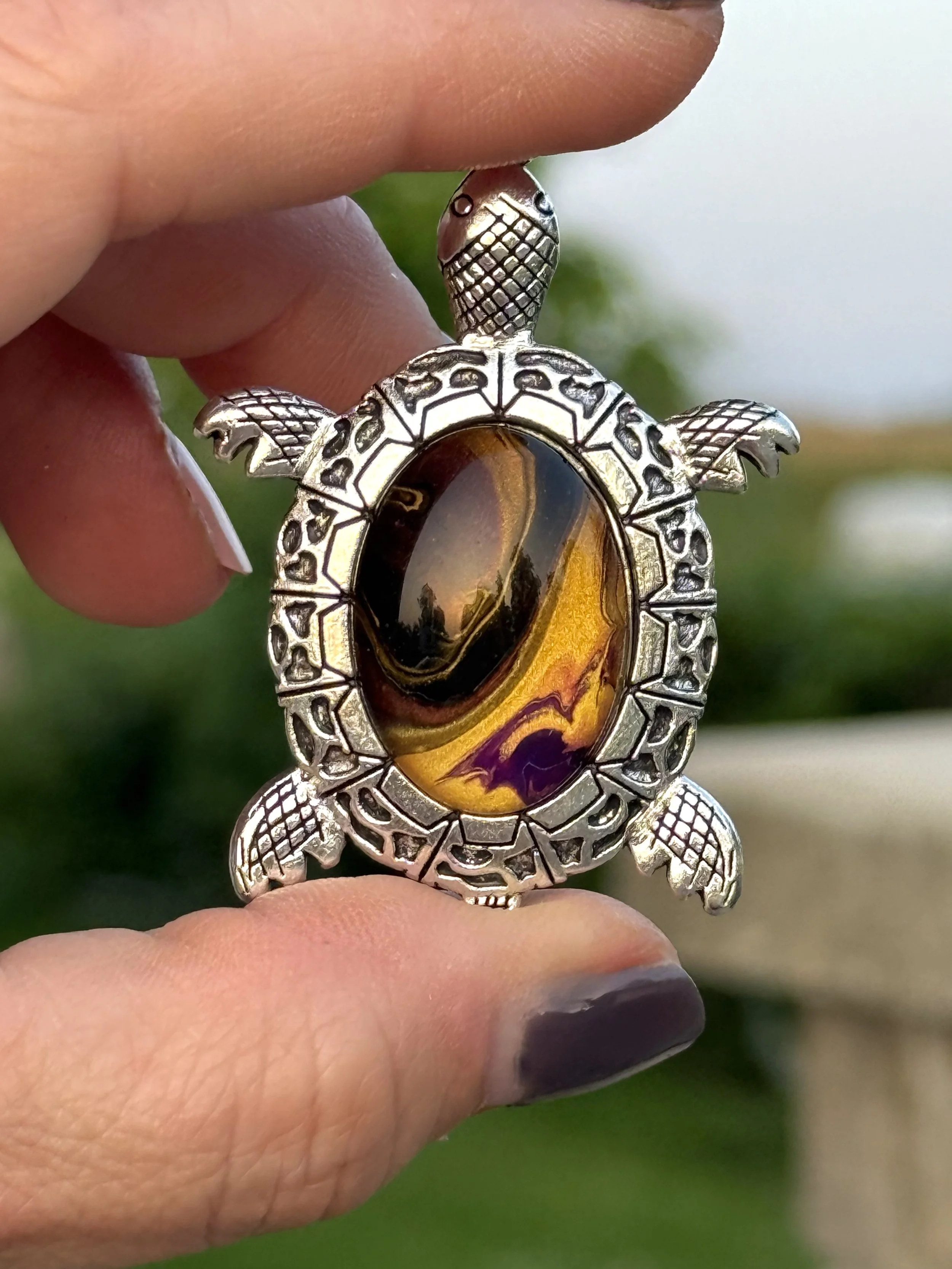 A close-up of a silver turtle-shaped pendant with an amber-colored gemstone in the center, held between fingers with dark nail polish, outdoors with blurred green background.