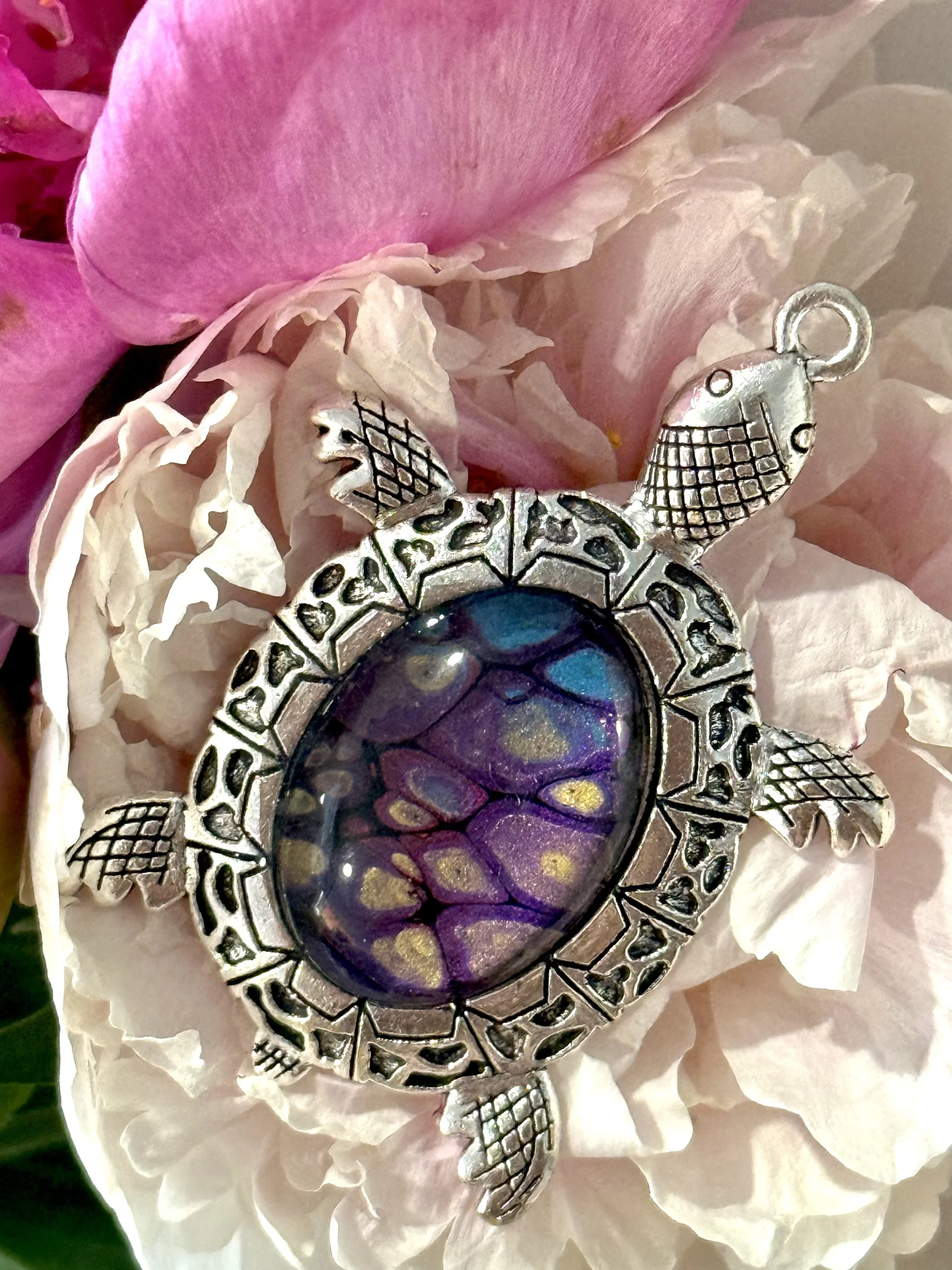 A close-up of a decorative turtle pendant with a dark purple, blue, and gold iridescent center, surrounded by white and pink flower petals.