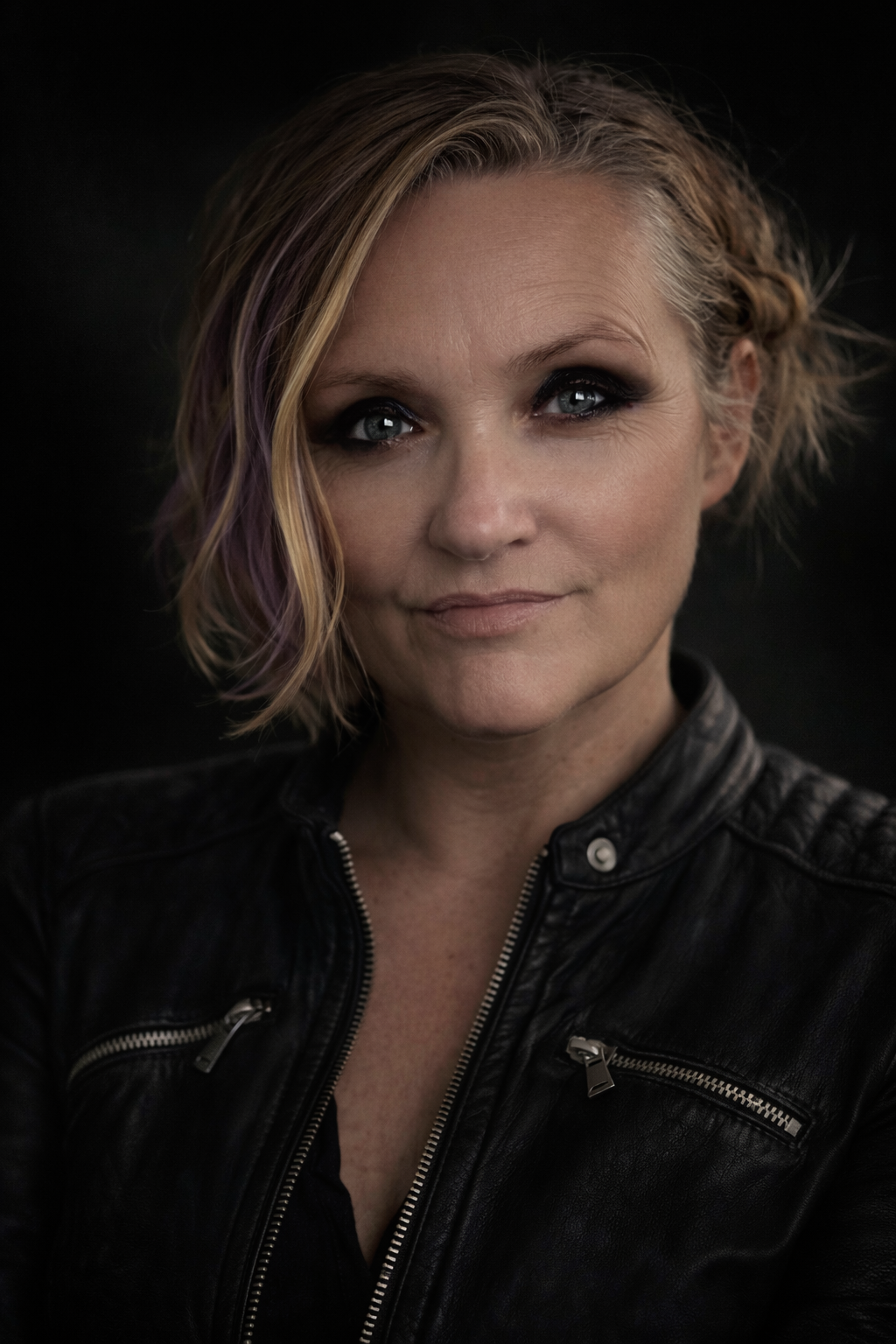Close-up portrait of a woman with short, wavy blonde hair with purple streaks, wearing a black leather jacket, against a dark background.