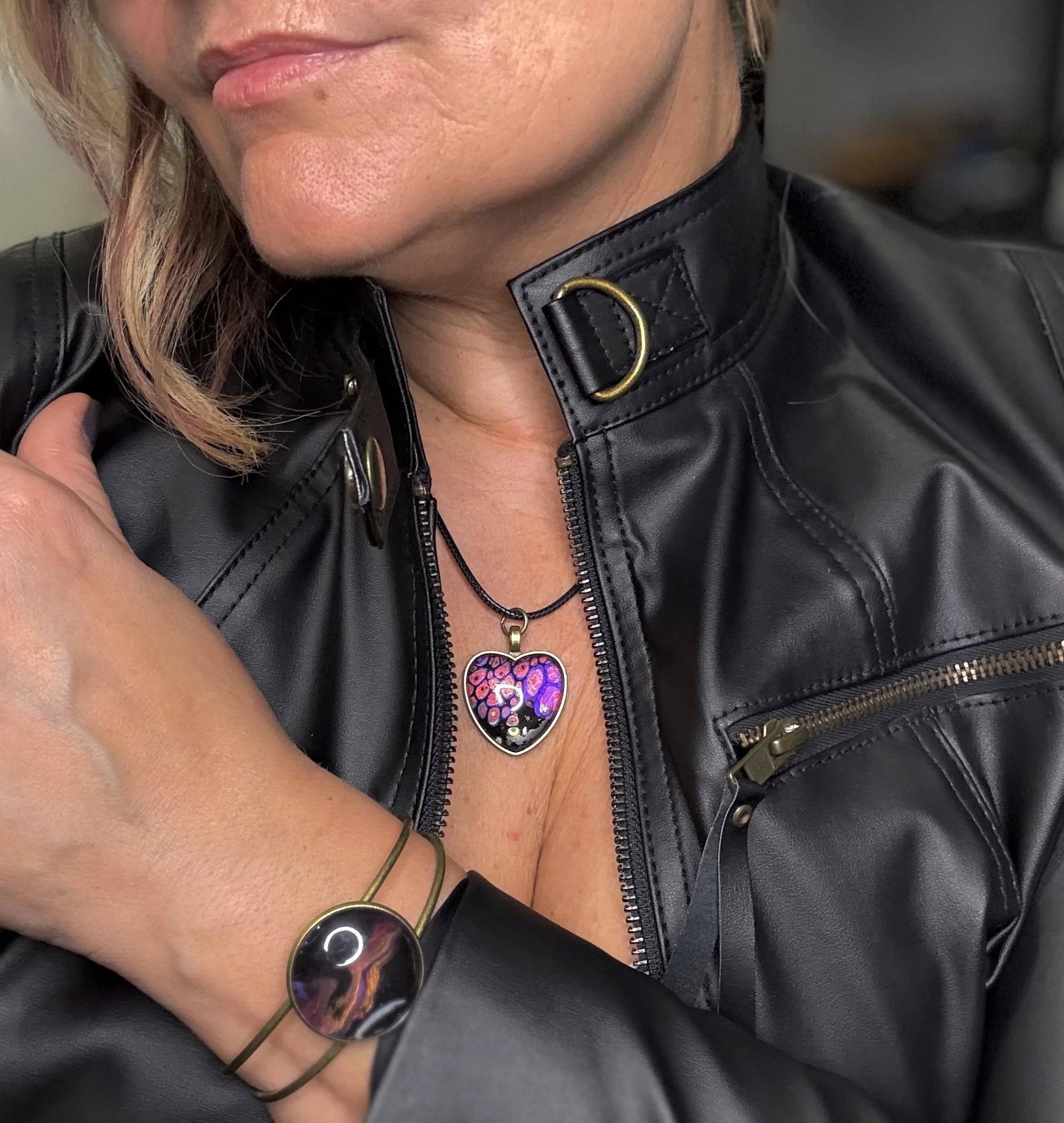 Close-up of a woman wearing a black leather jacket with a decorative buckle, holding sunglasses in front of her chest, and wearing a necklace with a heart-shaped pendant. She has light brown hair and tan skin.