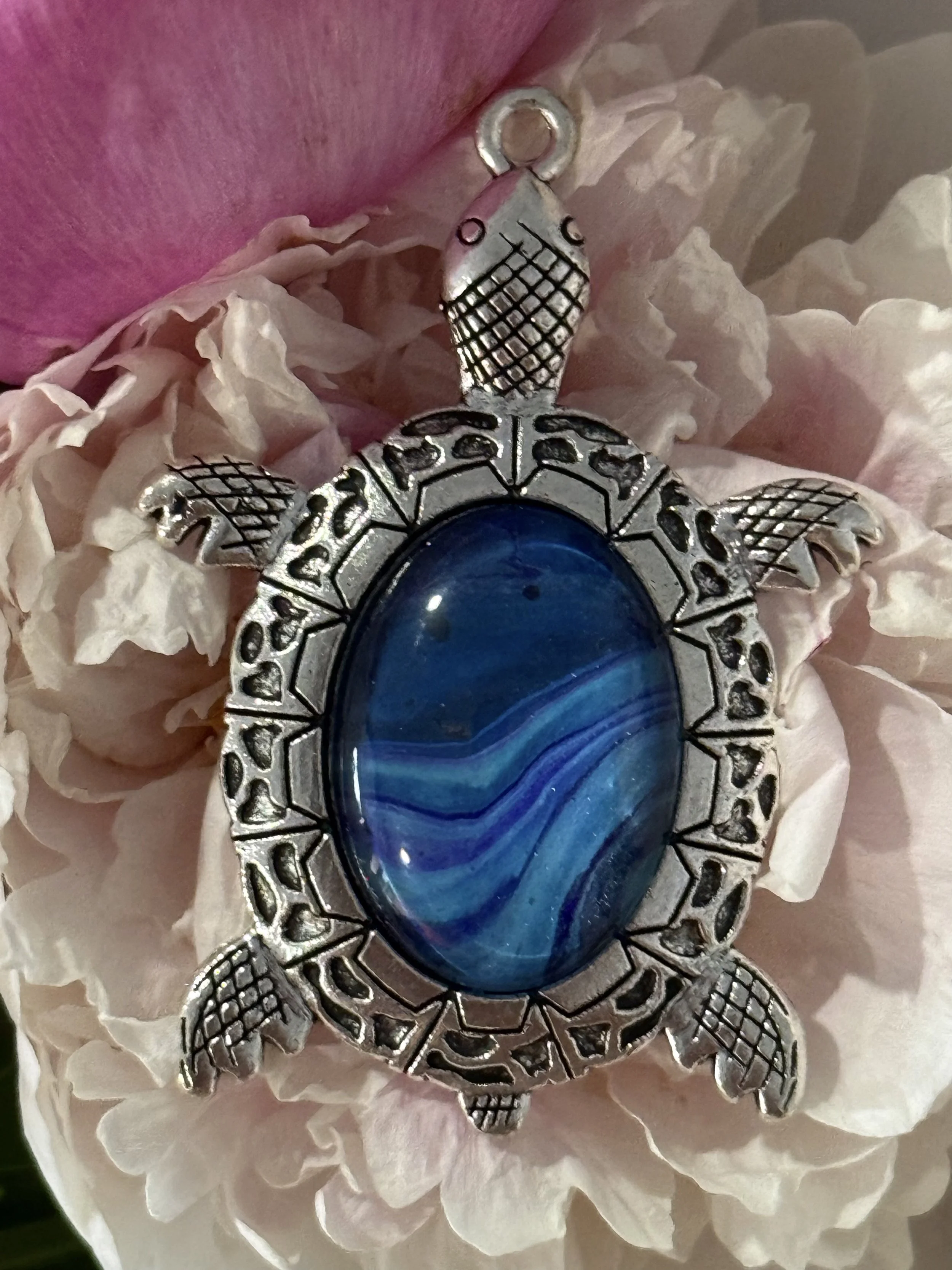 A silver turtle-shaped pendant with a large blue oval gemstone in its center, surrounded by white petals and pink flower petals.