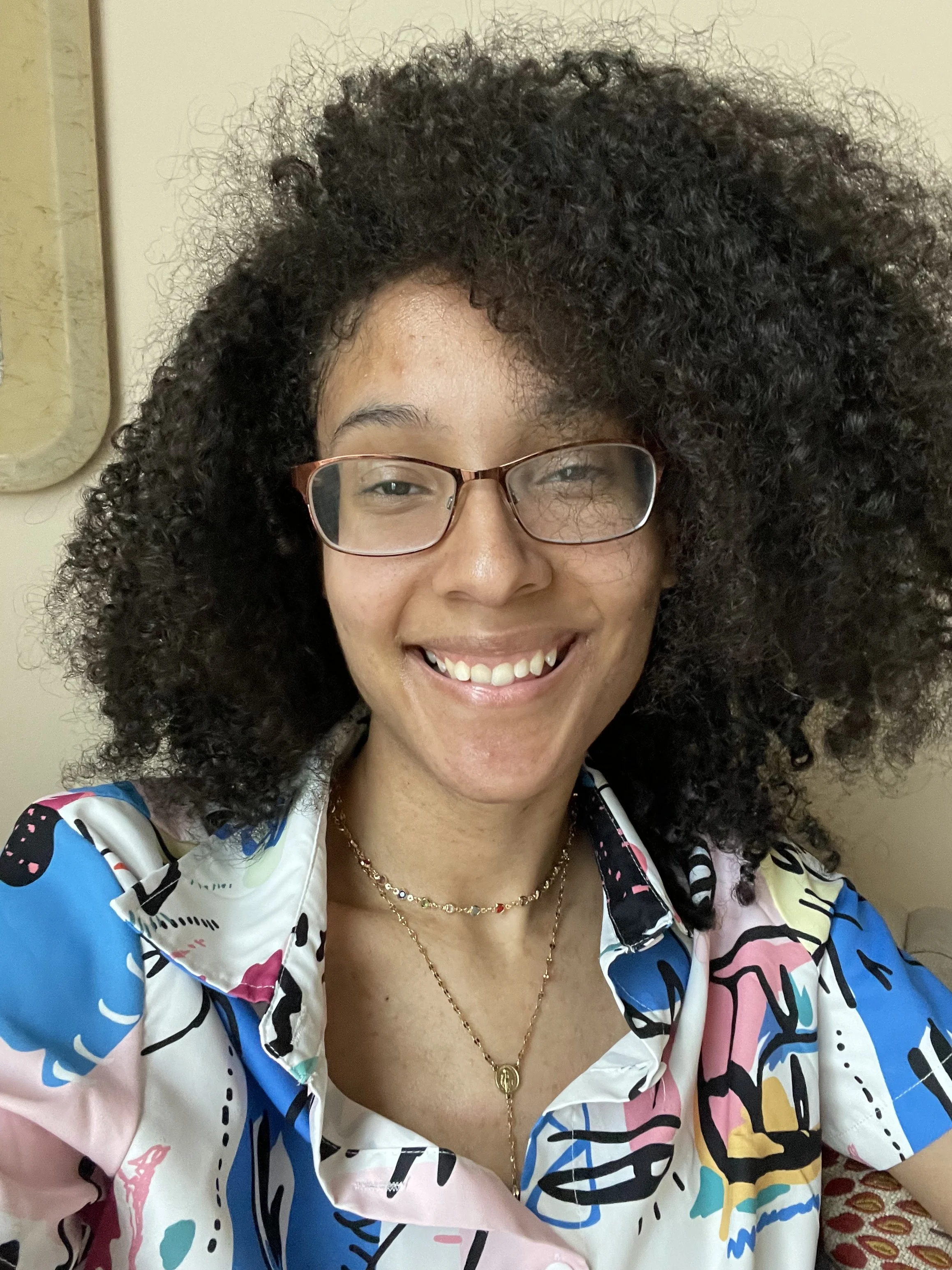 A woman with curly black hair, glasses, and a wide smile, wearing a colorful abstract-patterned shirt and layered necklaces, taking a selfie indoors.