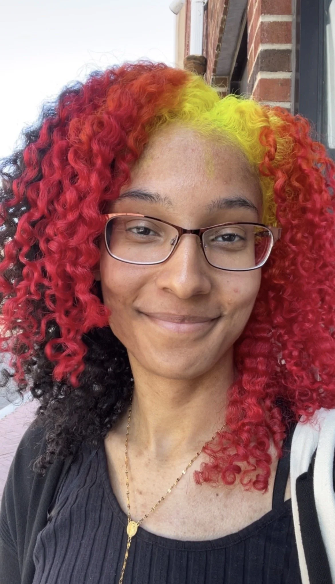 A woman with curly, brightly colored hair in red, yellow, and black, wearing glasses and a black top, smiling outdoors near a brick building.