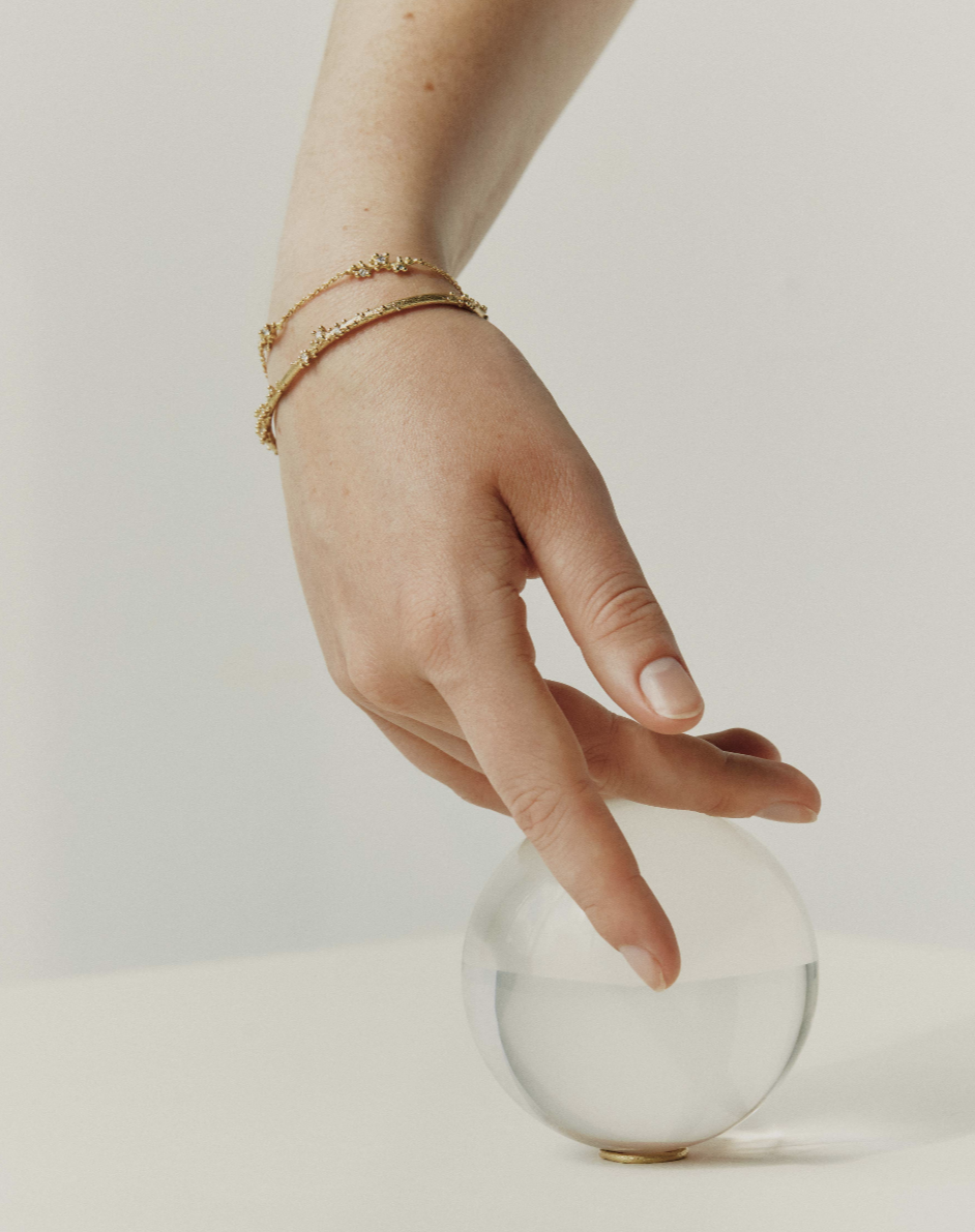 A hand with gold bracelets gently touching a round object with water inside, set against a plain white background.