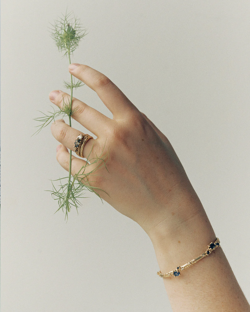 A hand with jewelry holding a sprig of green feathery plant against a neutral background.