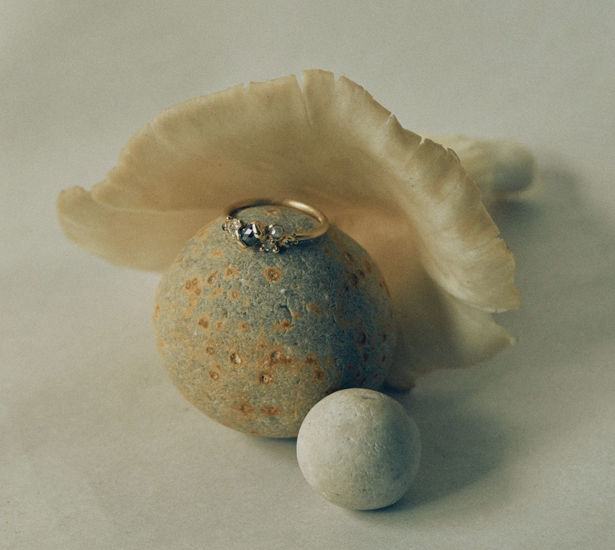 A gold ring with dark stones resting on a rounded, speckled stone, surrounded by a seashell and two smaller white spherical stones.