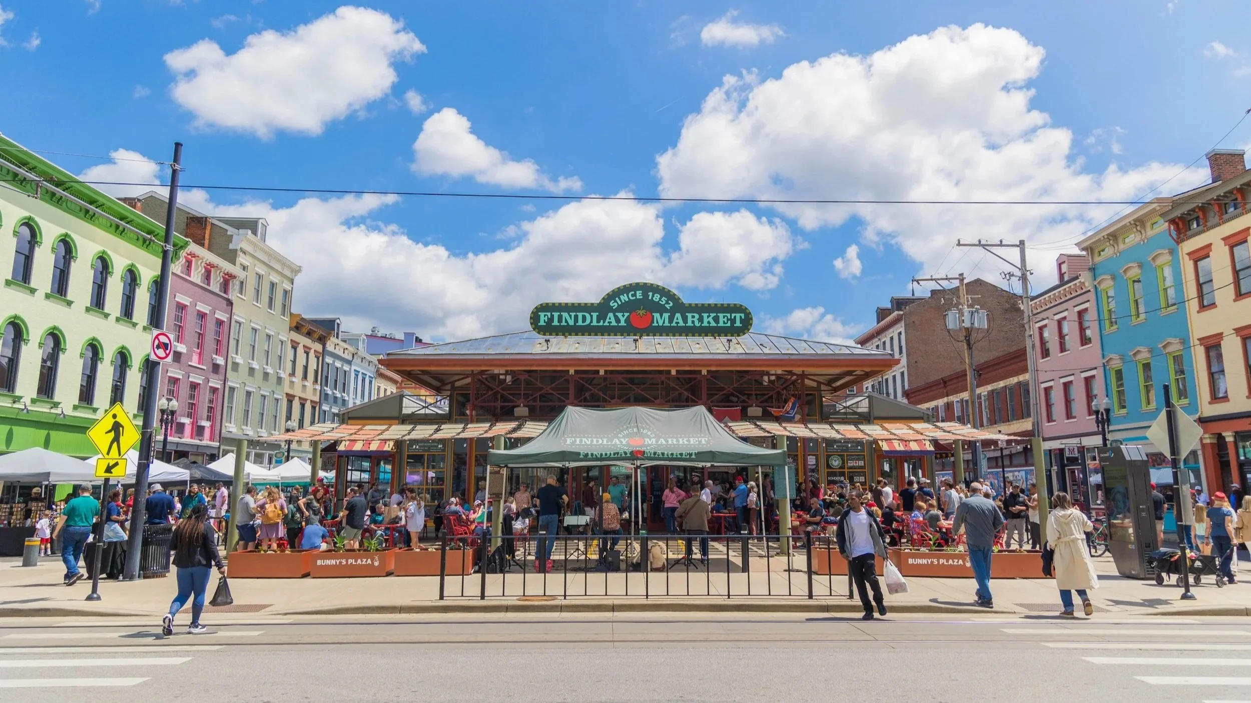 Crowd of people in front of Findlay Market with colorful buildings under a partly cloudy sky.