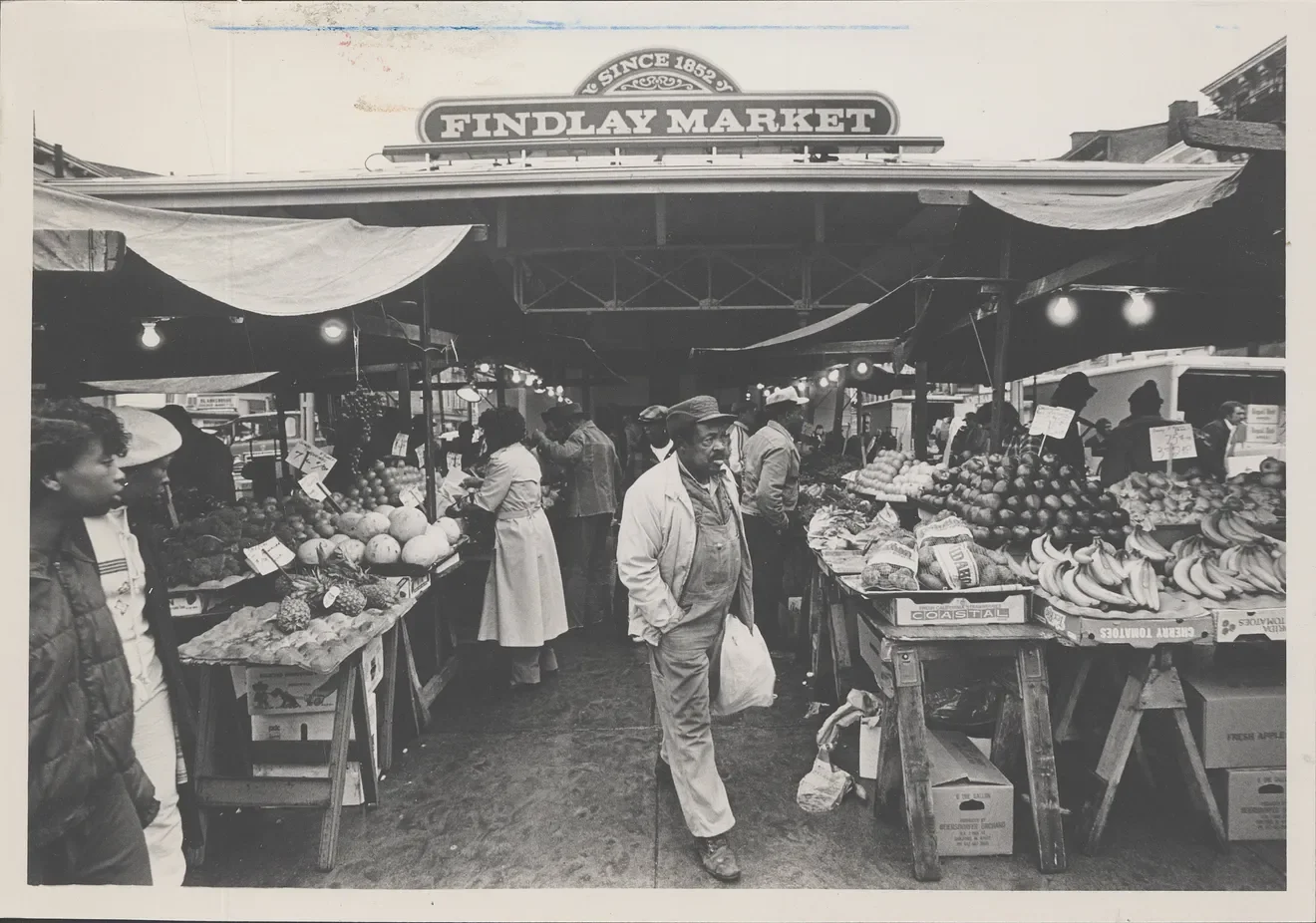 Black and white photo of a bustling outdoor farmers market with people shopping for fresh produce including fruits and vegetables