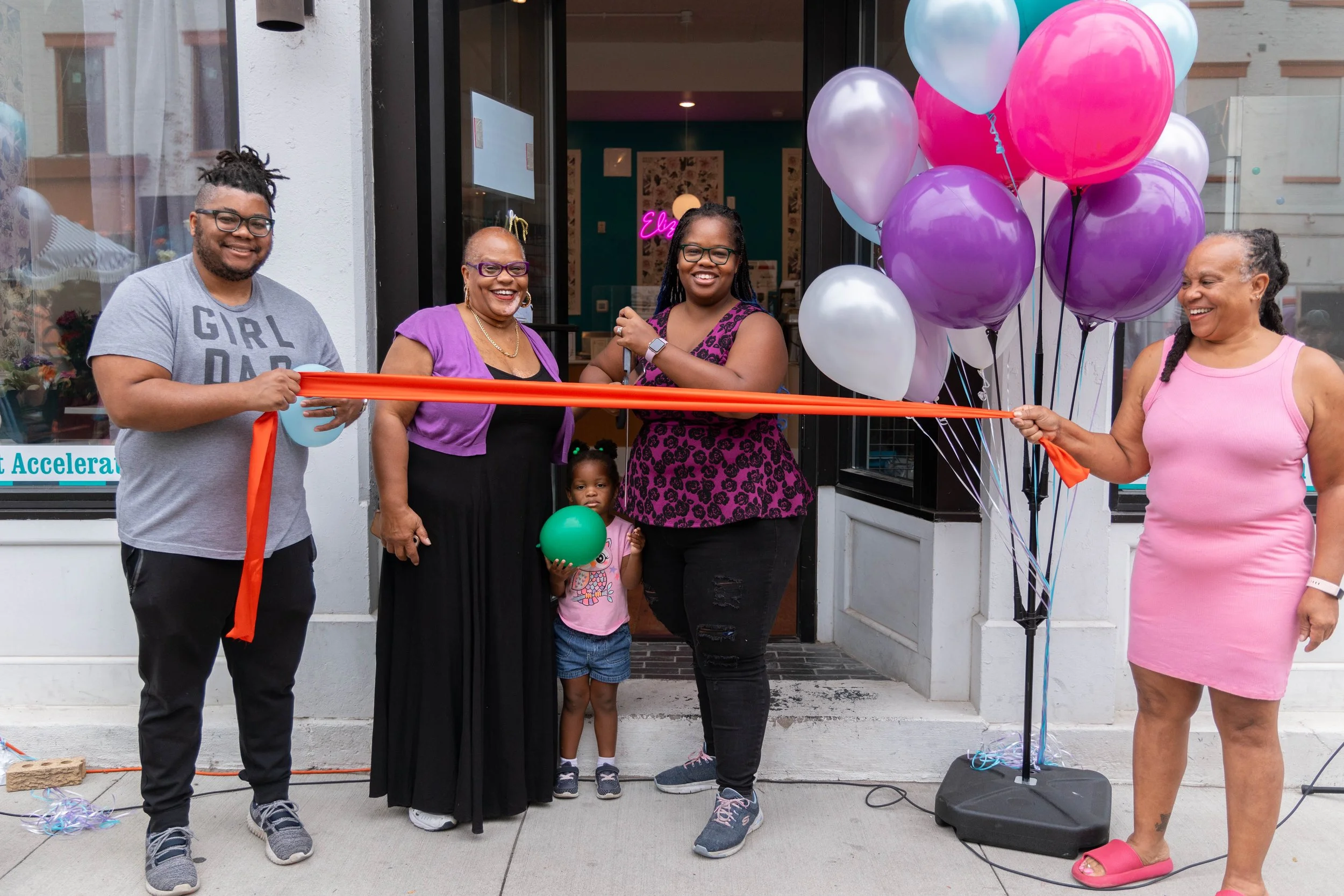 A group of six women and two children standing in front of a building during a ribbon-cutting event, with balloons and a microphone stand nearby.
