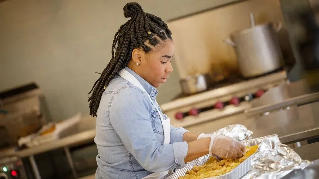 Young woman with braided hair wearing gloves and an apron, preparing food in a kitchen with stoves and large pots in the background.
