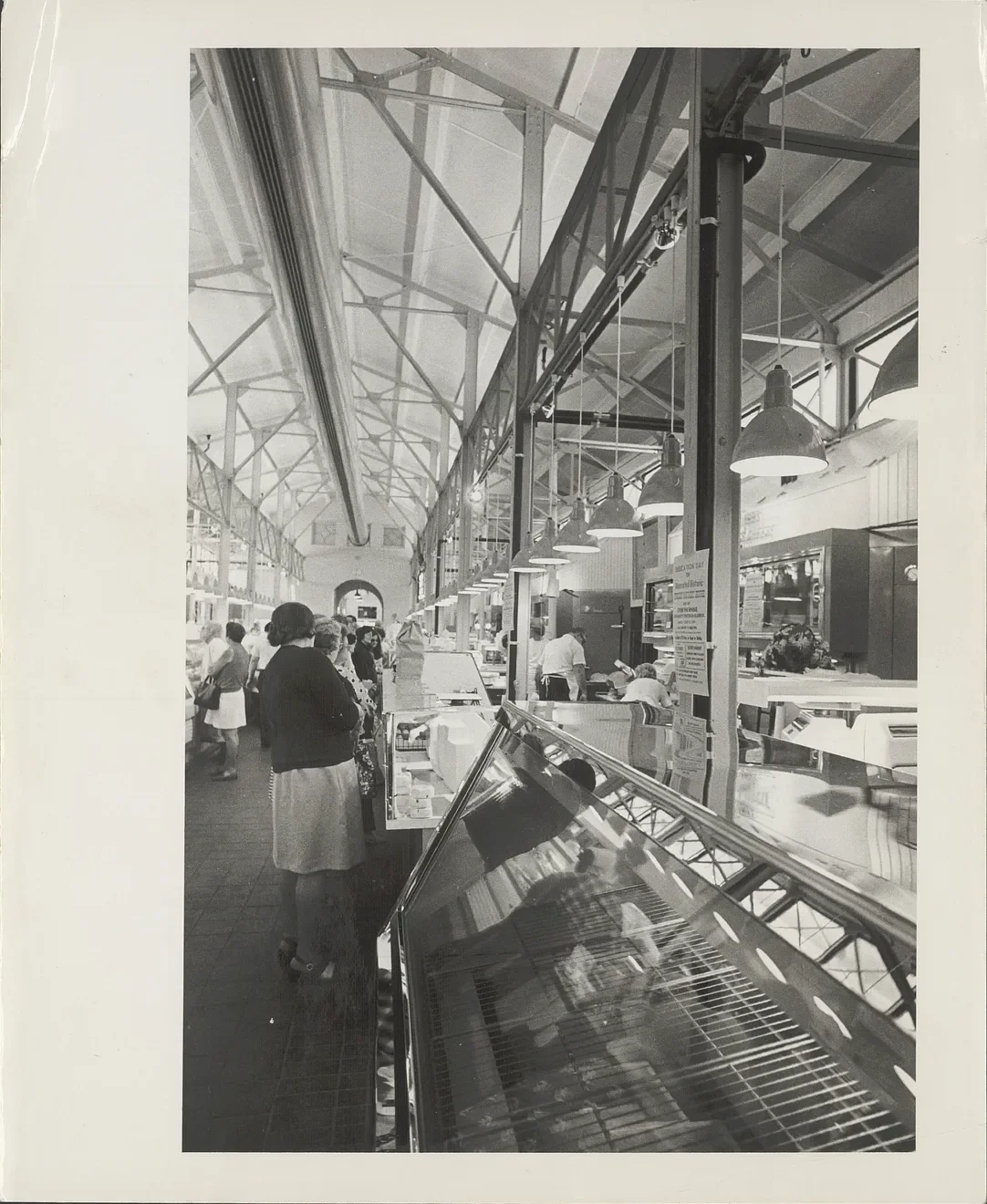 Black and white photo of a marketplace or indoor food stall with people shopping and working behind counters, featuring a high ceiling with metal trusses and hanging industrial lights.