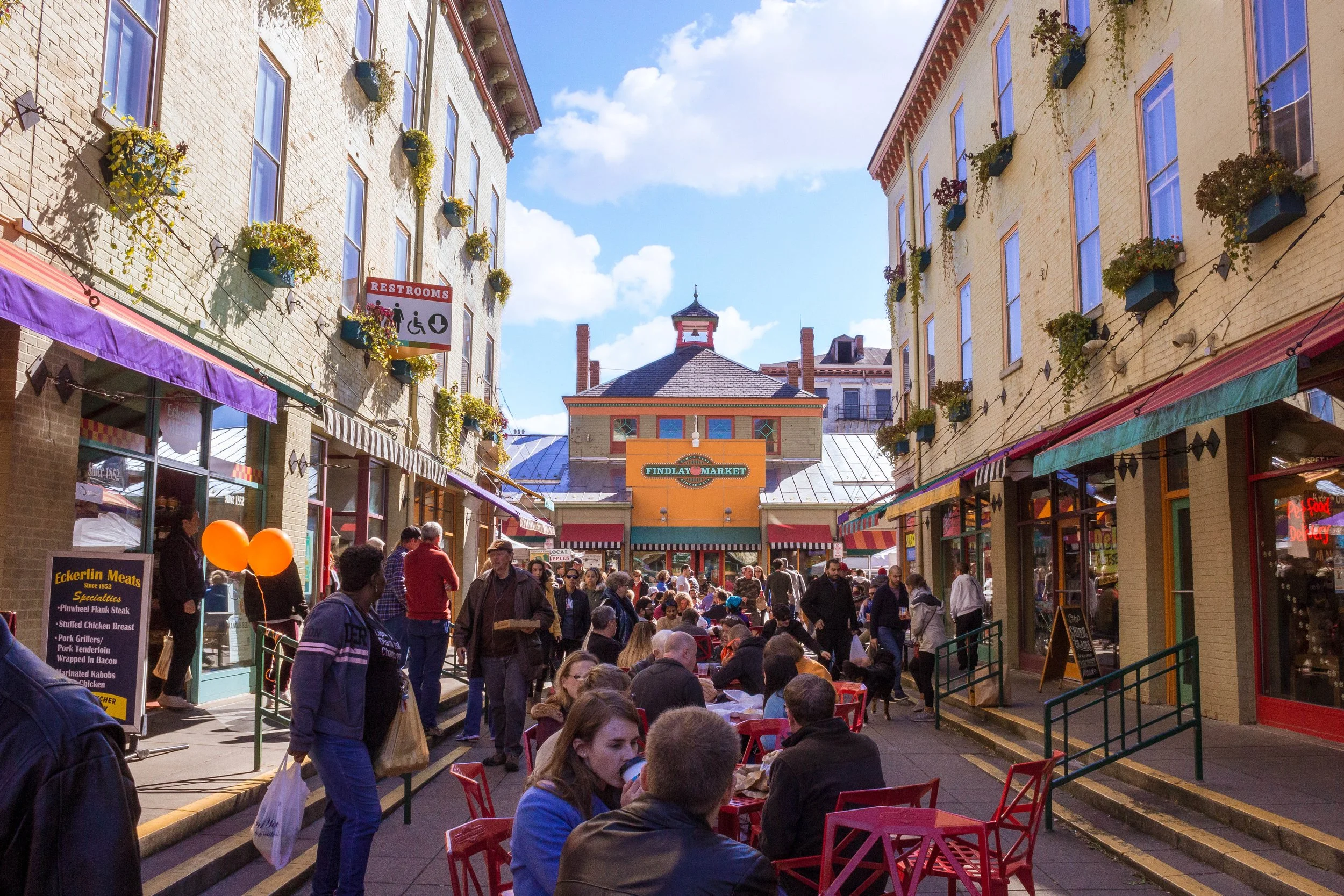A bustling outdoor market street with outdoor seating, colorful umbrellas, shop signs, and many people walking and sitting, under clear blue skies.