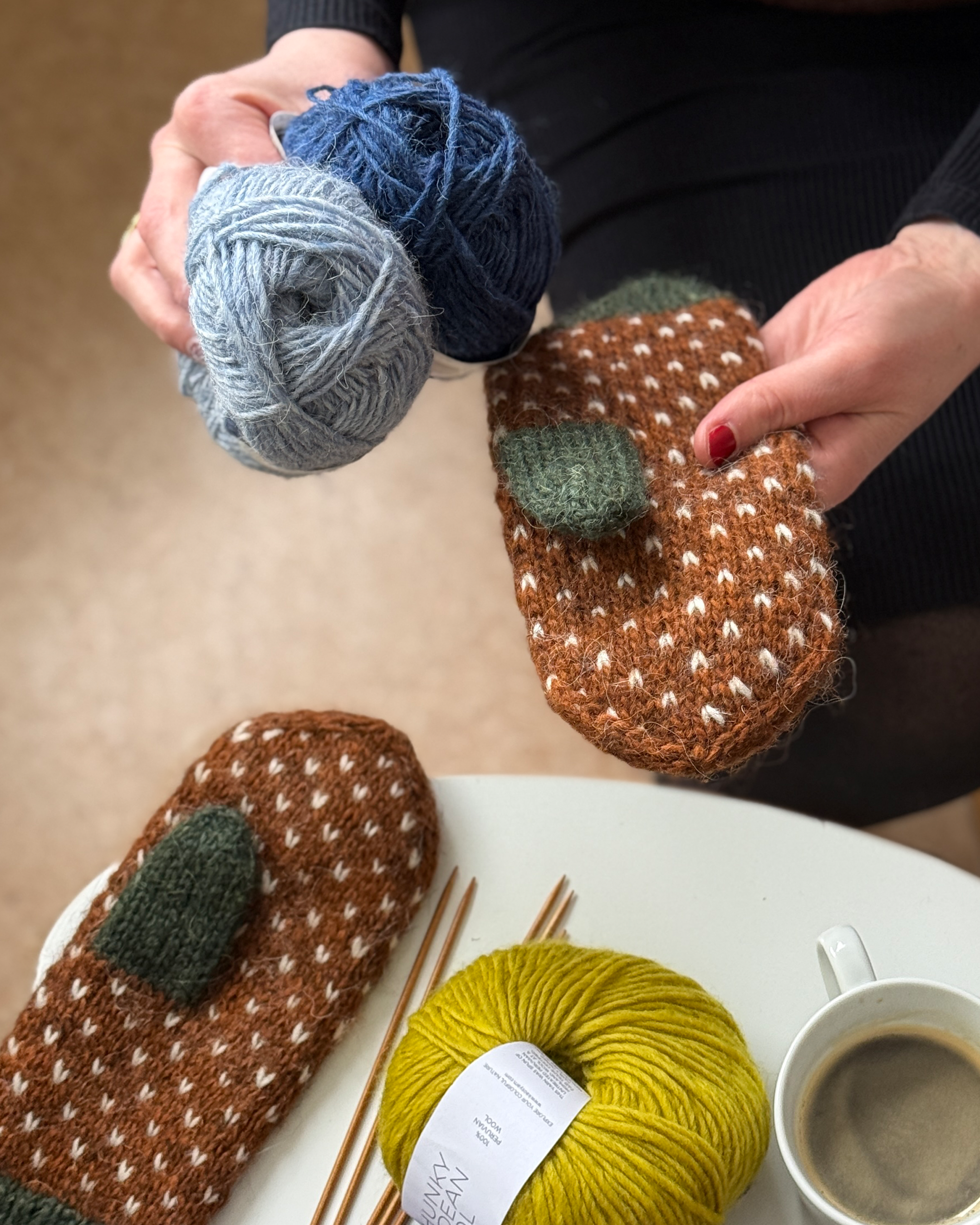 Person holding a knitted mitten with wool yarn in shades of blue and gray, and a partially completed mitten with green and brown yarn on a white surface with a cup of coffee and knitting needles nearby.