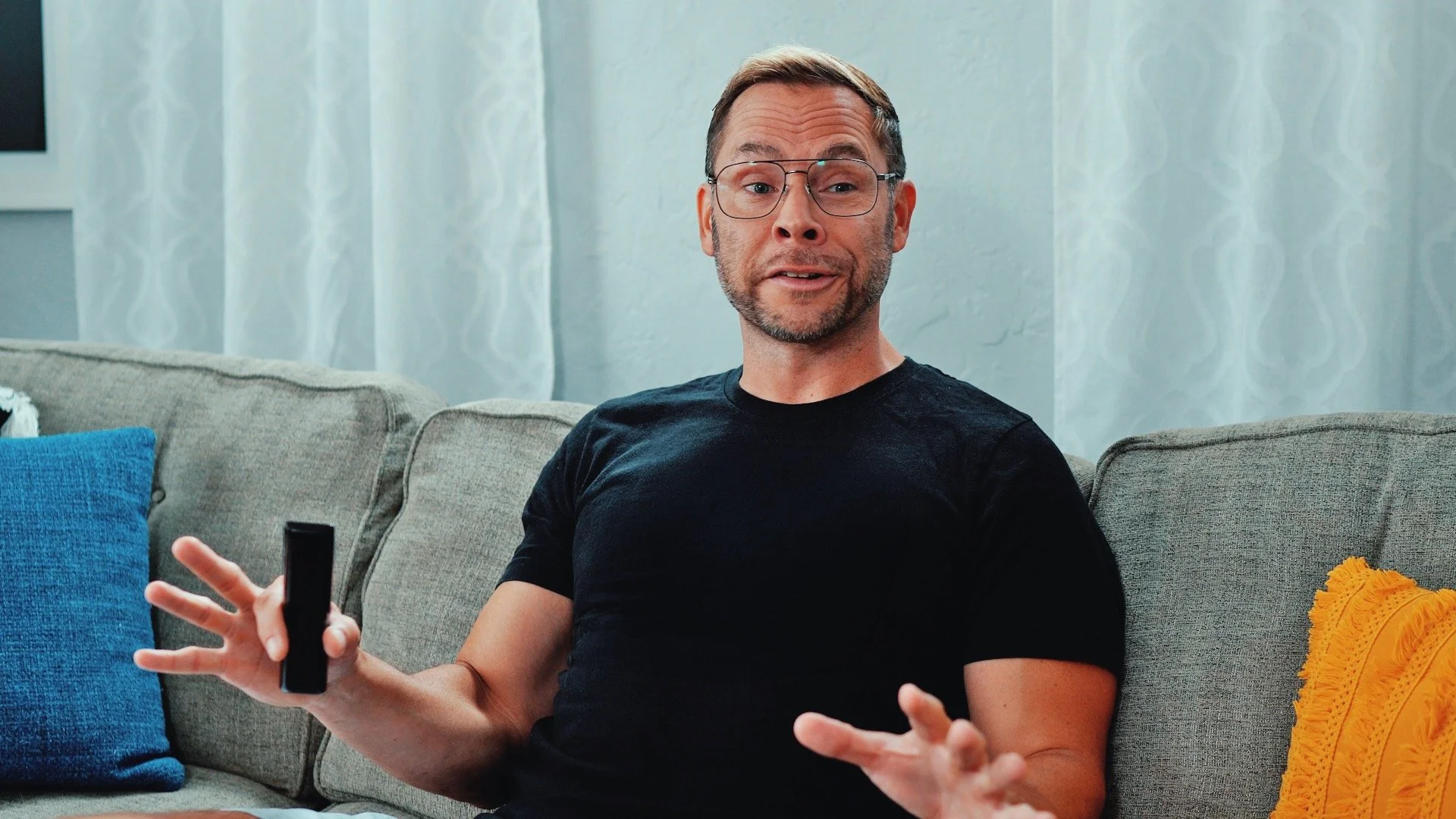 A man with glasses and a beard, wearing a black t-shirt, sits on a beige couch with colorful pillows, holding a TV remote, and gesturing with his hands, in a living room with light blue walls and patterned curtains.