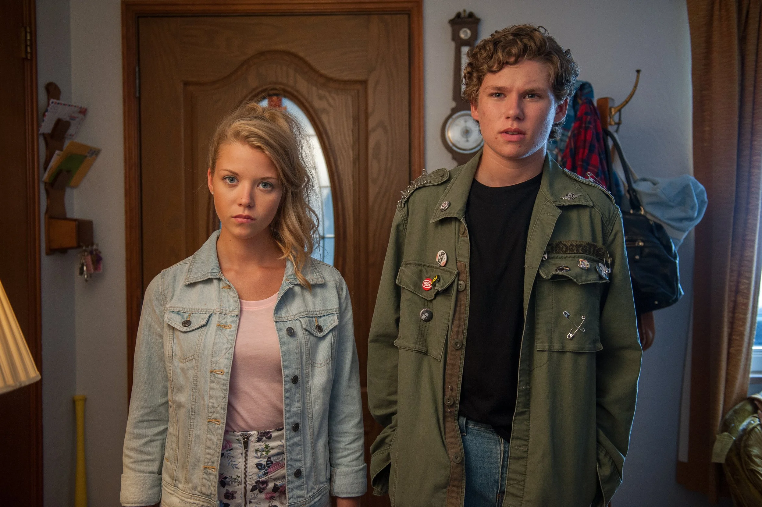 A young woman and a young man standing in a hallway, looking toward the camera. The woman has blonde hair, blue eyes, and is wearing a denim jacket over a pink top with floral pants. The man has curly hair and is wearing a green military-style jacket