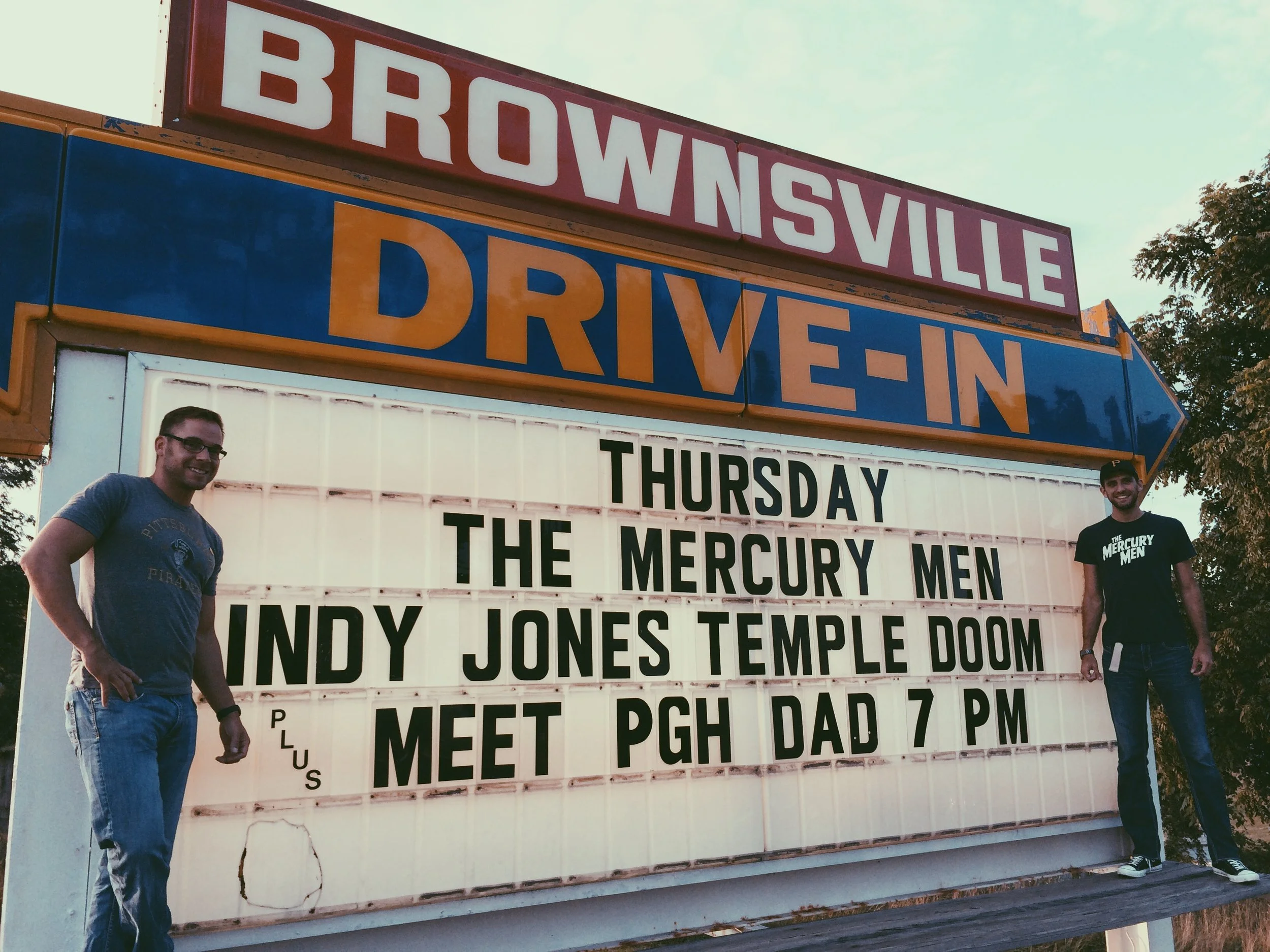 Pittsburgh Dad star Curt Wootton and director Chris Preksta posing in front of drive-in movie theater sign advertising a screening of The Mercury Men, Indiana Jones and the Temple of Doom, and Pittsburgh Dad.