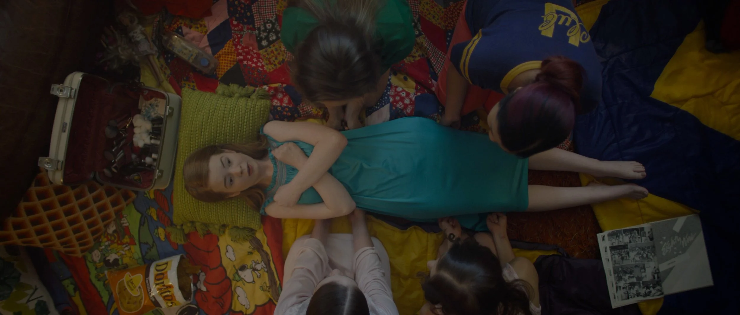 Doll laying on blanket surrounded by children and women, with makeup items and snacks nearby.