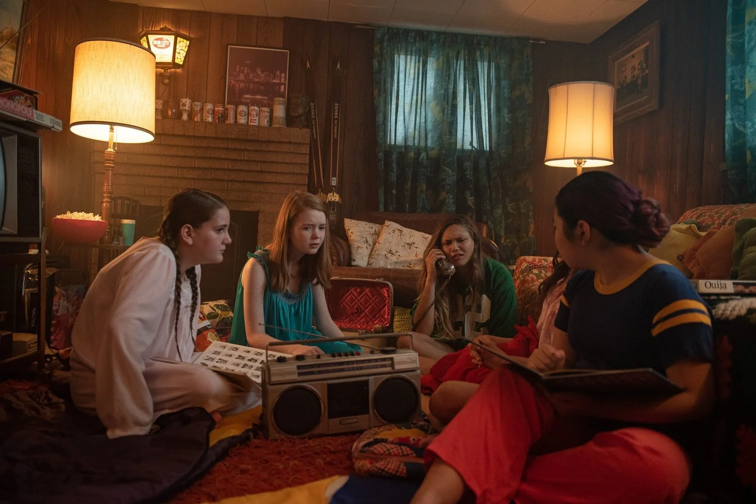 Four young women sitting on the floor of a cozy living room, engaged in a conversation. The room has wooden paneled walls, lamps providing warm lighting, and a window with curtains. There are various objects like a boombox, puzzle, popcorn, and board