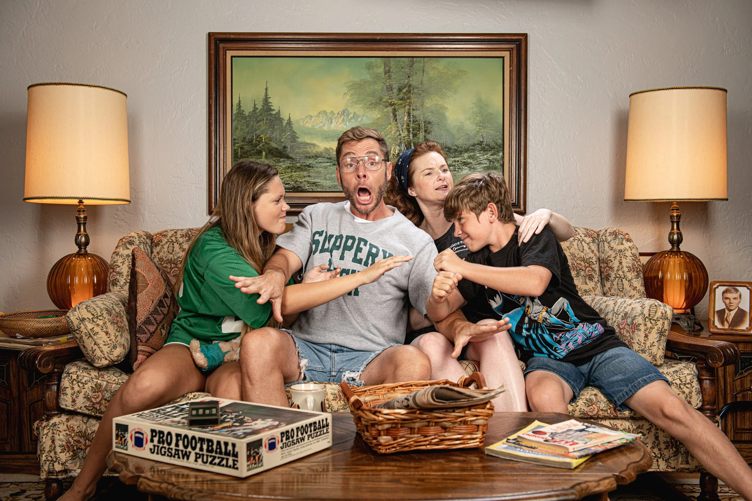 A family of five sitting on a vintage floral sofa in a living room, engaging in a lively discussion. The room features two table lamps, a framed nature painting on the wall, a framed photograph on the side table, and a coffee table with magazines and a boxed puzzle labeled "Pro Football Jigsaw Puzzle."