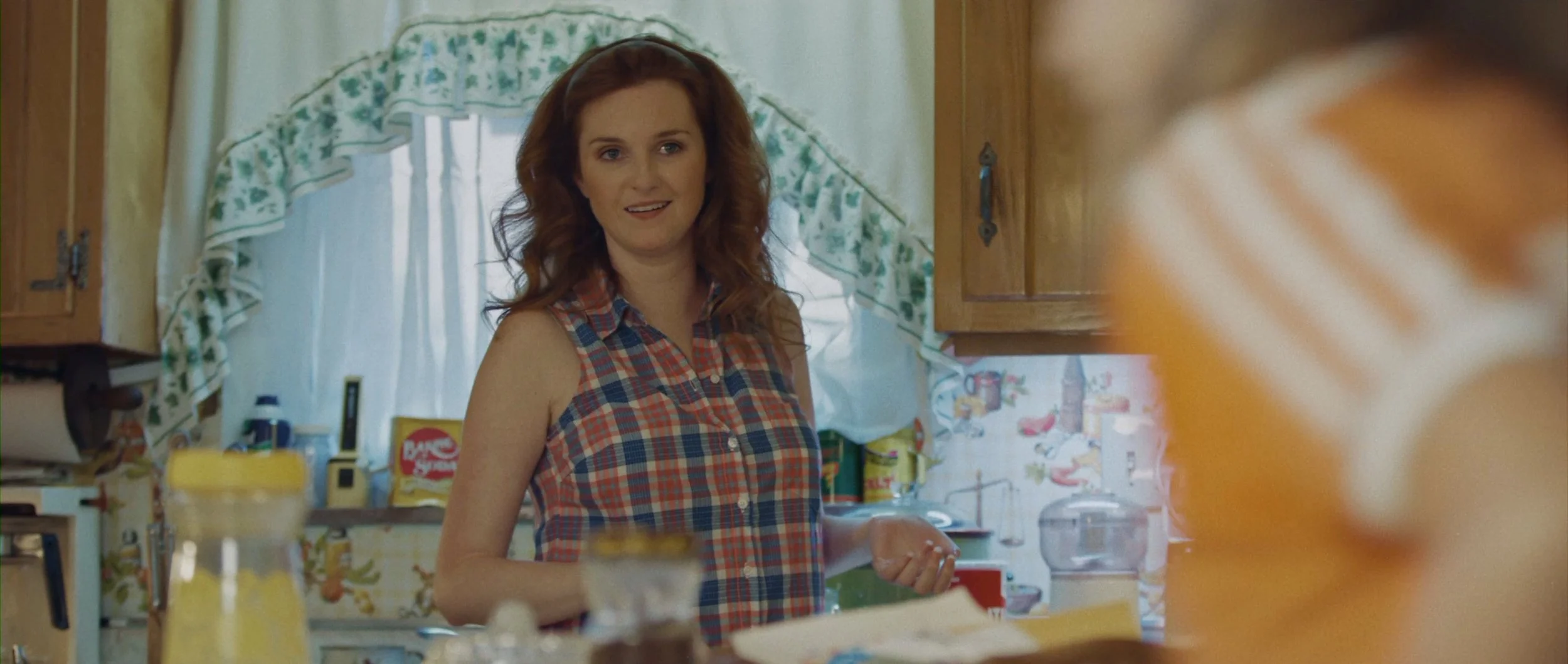 A woman with red hair wearing a sleeveless plaid shirt standing in a kitchen.