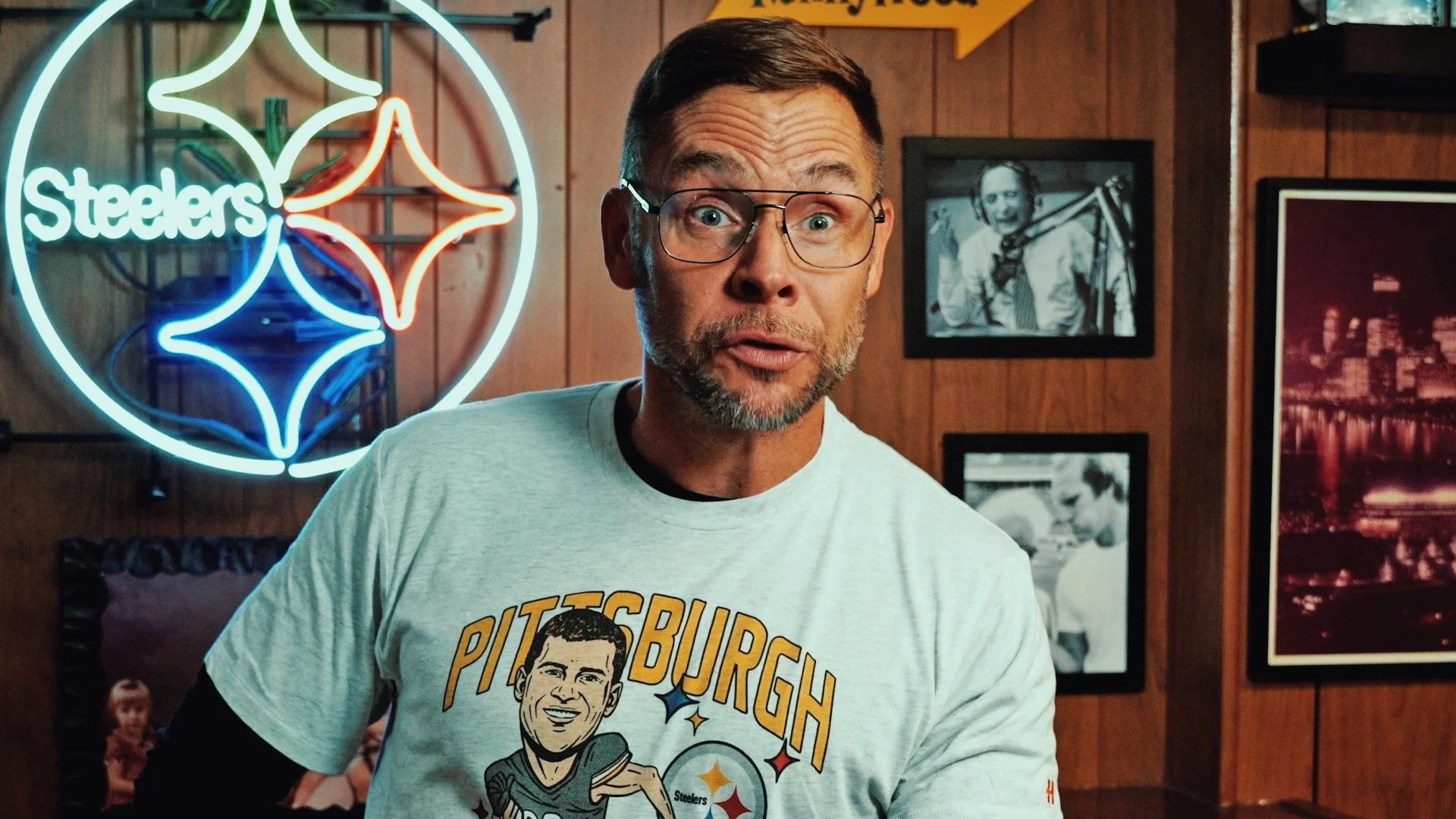 A man wearing glasses and a Pittsburgh Steelers T-shirt is standing in a bar or restaurant. Behind him, there is a neon Steelers sign and framed photos on a wood-paneled wall.