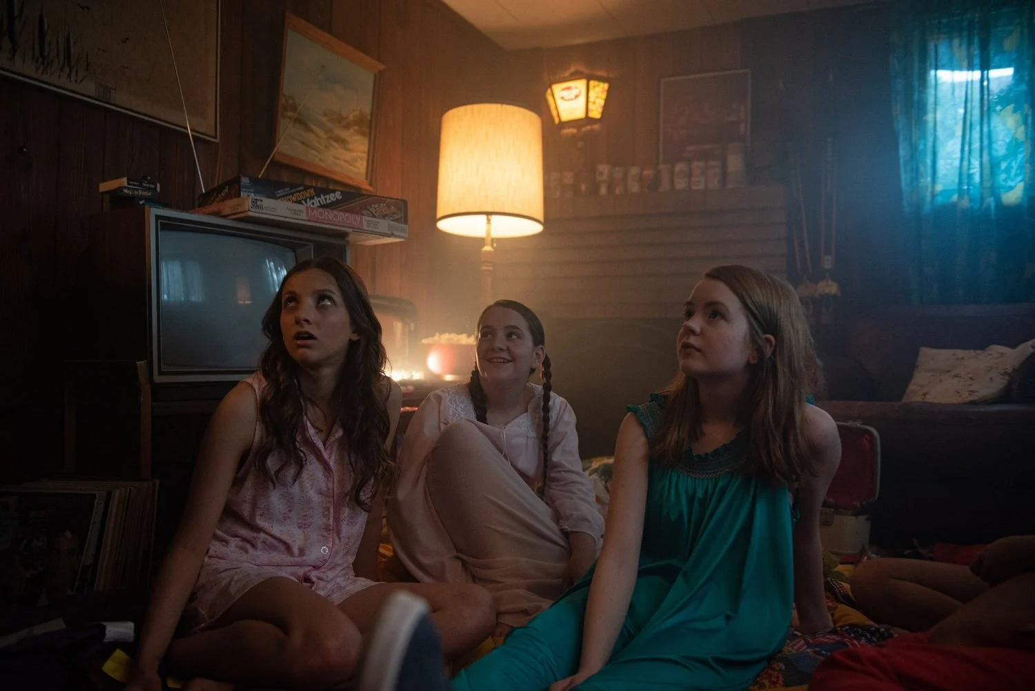 Three young girls sitting on the floor in a dimly lit room, watching television with expressions of surprise and interest.