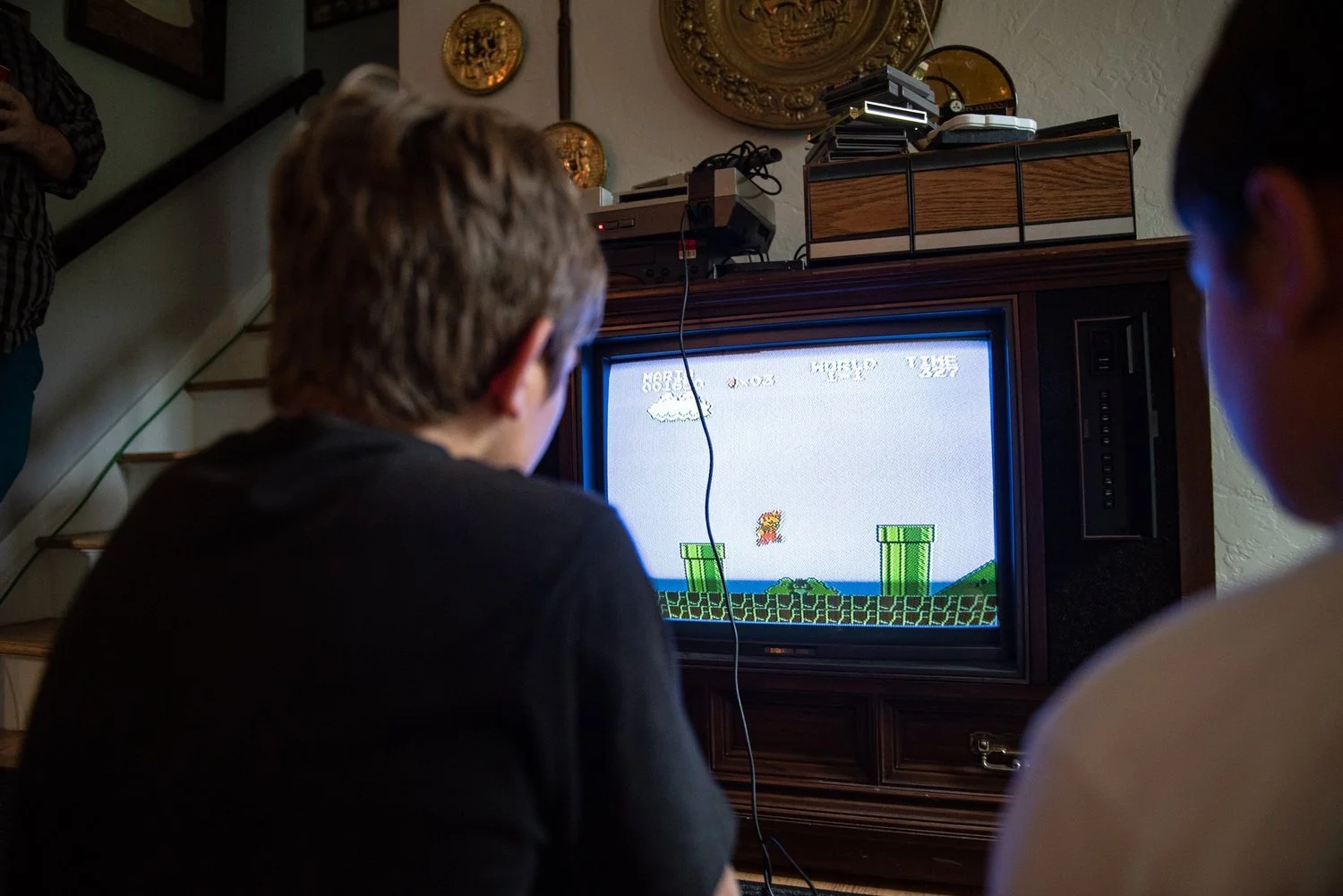 Two boys are sitting in front of a TV playing Super Mario Bros. on an old school gaming console. They are in a living room with decorative plates hanging on the wall.