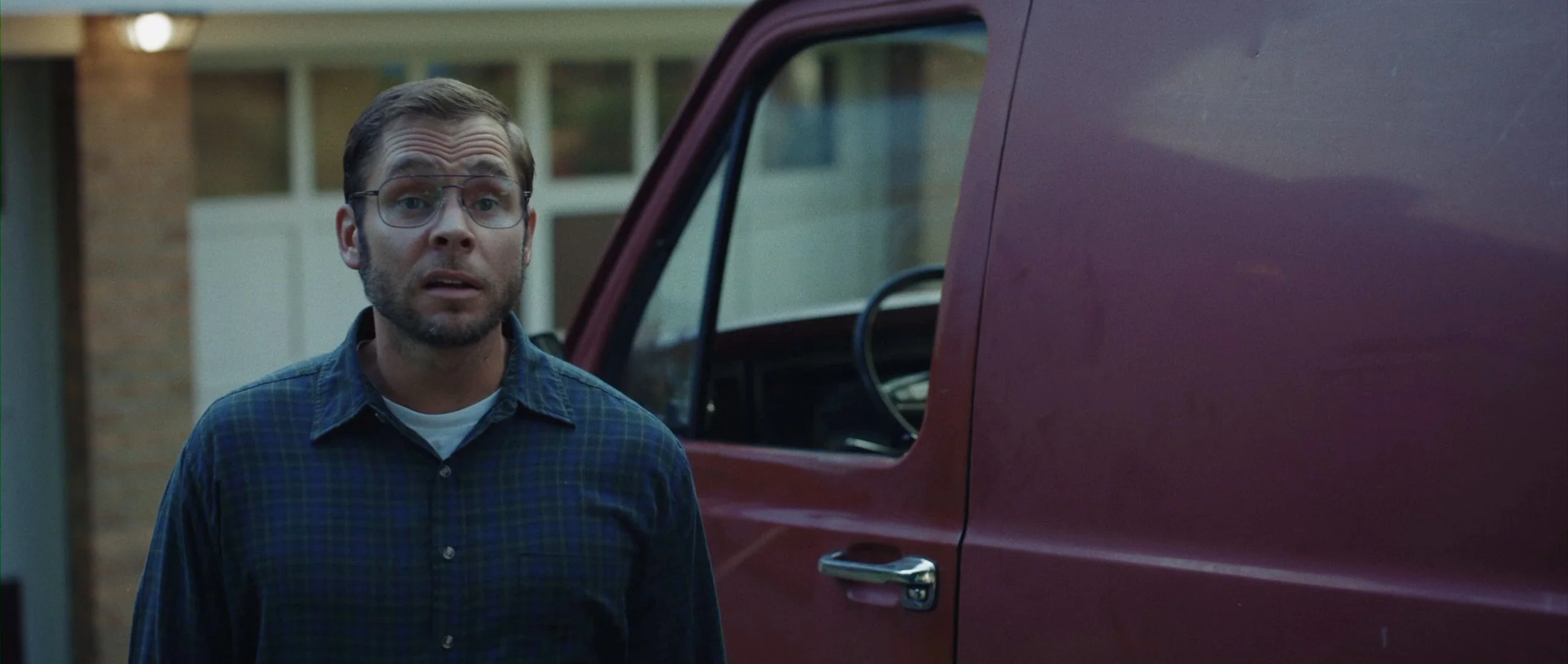 A man with glasses and a beard, wearing a blue checkered shirt, stands outdoors next to a red vehicle with an open door, in front of a suburban house.