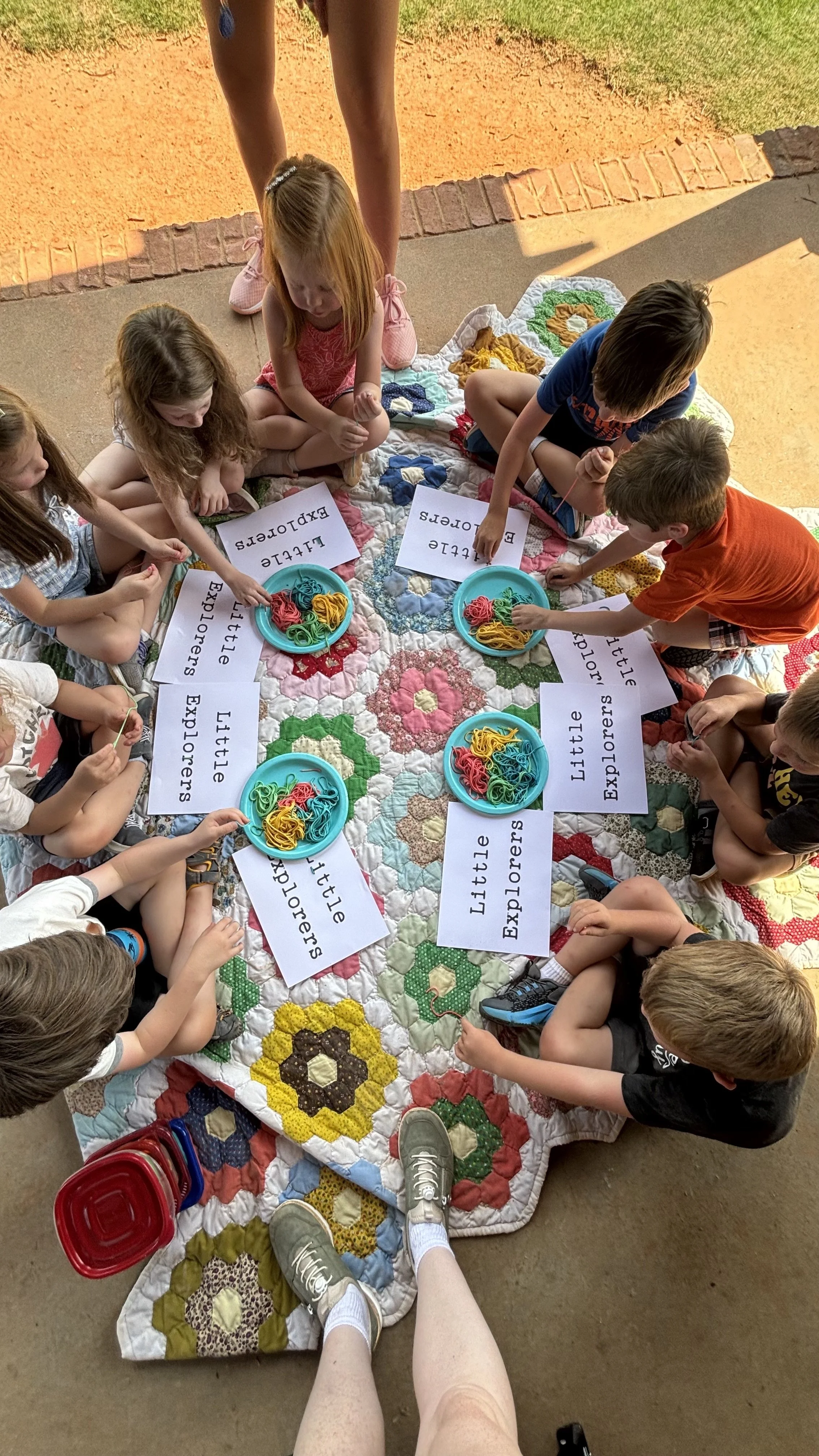 Children sitting on a quilt on the ground, sorting colorful rubber bands into bowls, with labels reading 'Little Explorers'.