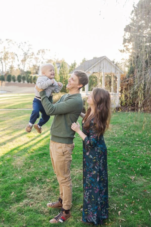 Man holding a smiling young child up in the air with a woman standing beside them in a backyard with a gazebo and trees.