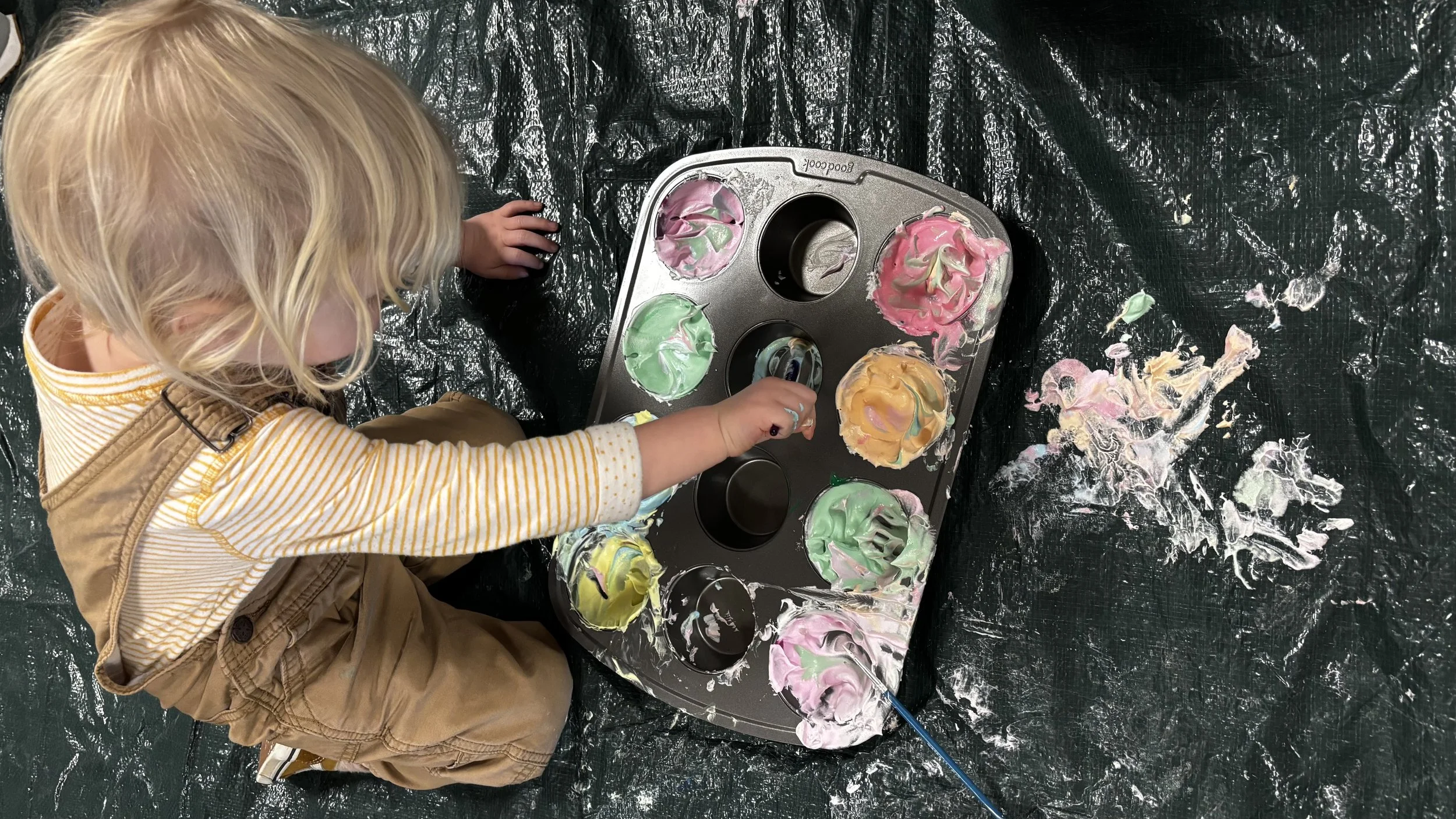 Child with blonde hair wearing a tan vest and yellow and white striped shirt painting with colorful paints on a black surface, with a baking tray filled with swirled paint in pink, green, yellow, and other colors.