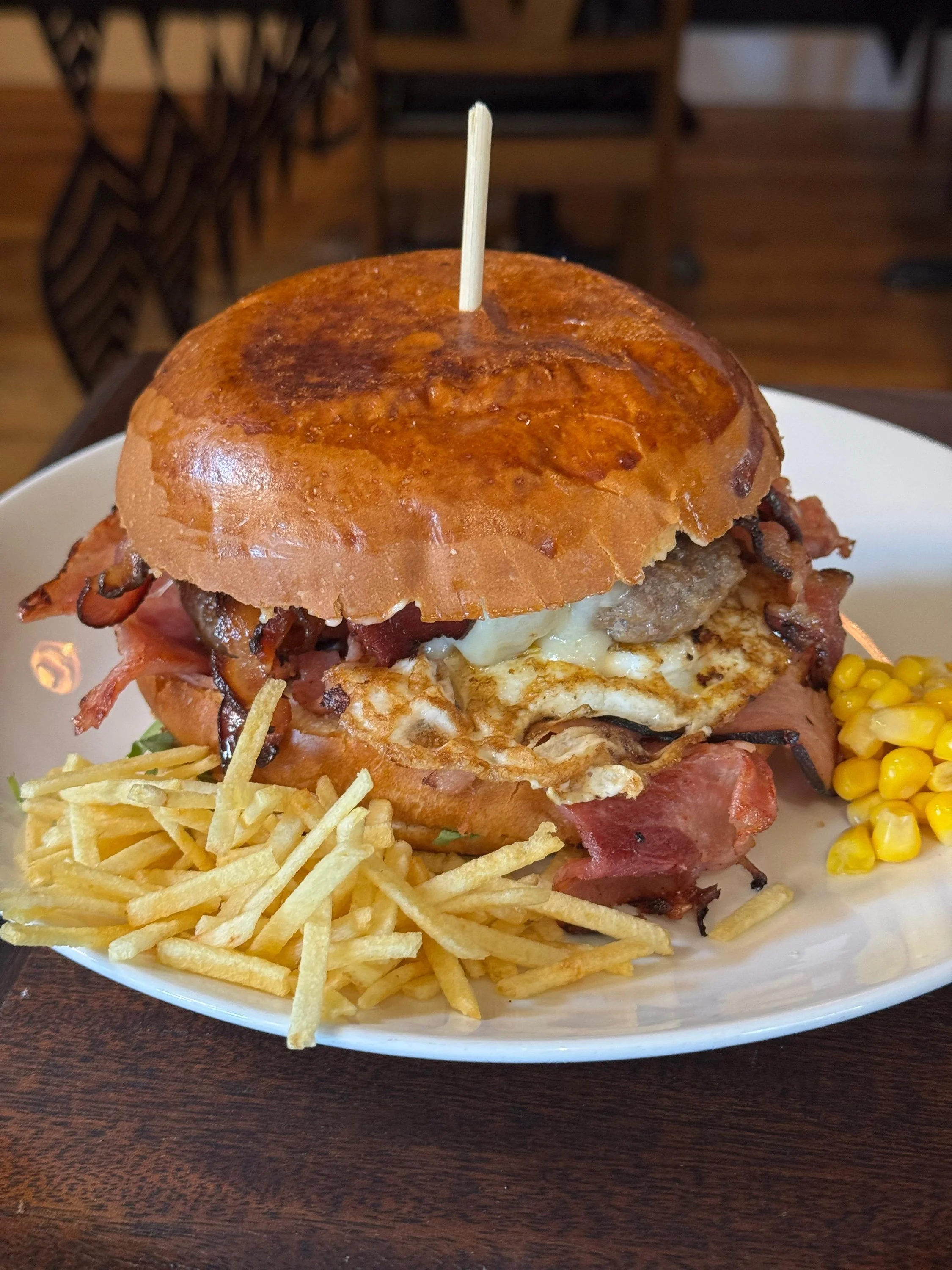 A burger with a bun top, crispy bacon, a fried egg, melted cheese, beef patty, and lettuce, served with a side of French fries and corn on a white plate.