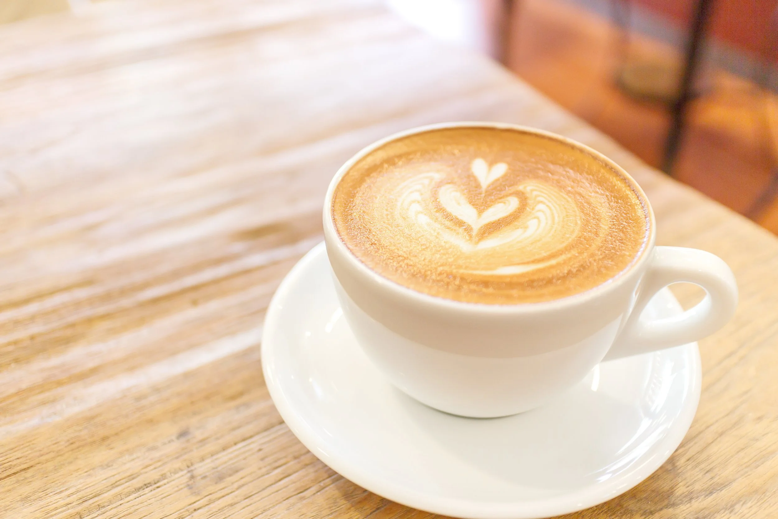 A cup of latte coffee with latte art on top, placed on a white saucer on a wooden table.